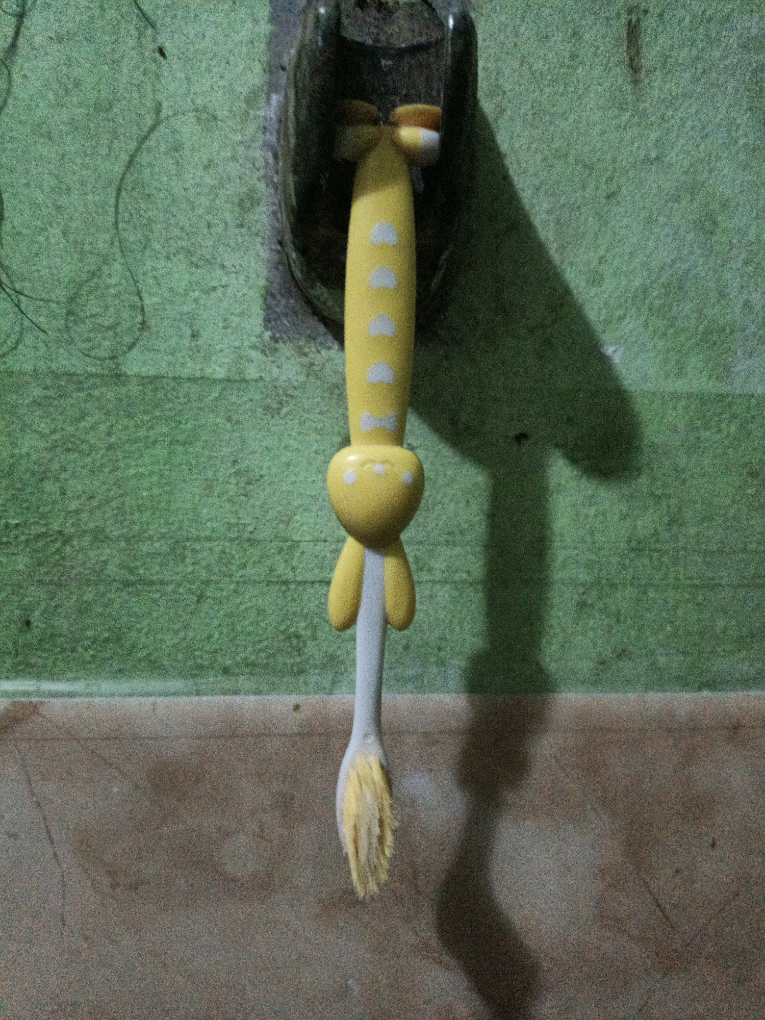 The image shows a yellow toothbrush with white dots, shaped like a giraffe, hanging from a holder. The toothbrush is positioned vertically, with its bristles pointing downwards. The background is a green, textured wall. A shadow of the toothbrush is cast on the wall to its right. The lower portion of the image shows a floor with a speckled pattern, appearing to be made of tile. The lighting suggests it might be nighttime or an indoor setting with artificial light. The overall scene is likely a bathroom or a similar domestic space. There is no visible text in the image. The image appears to be taken in West Jakarta, Indonesia, though this context does not provide visual cues within the image itself.