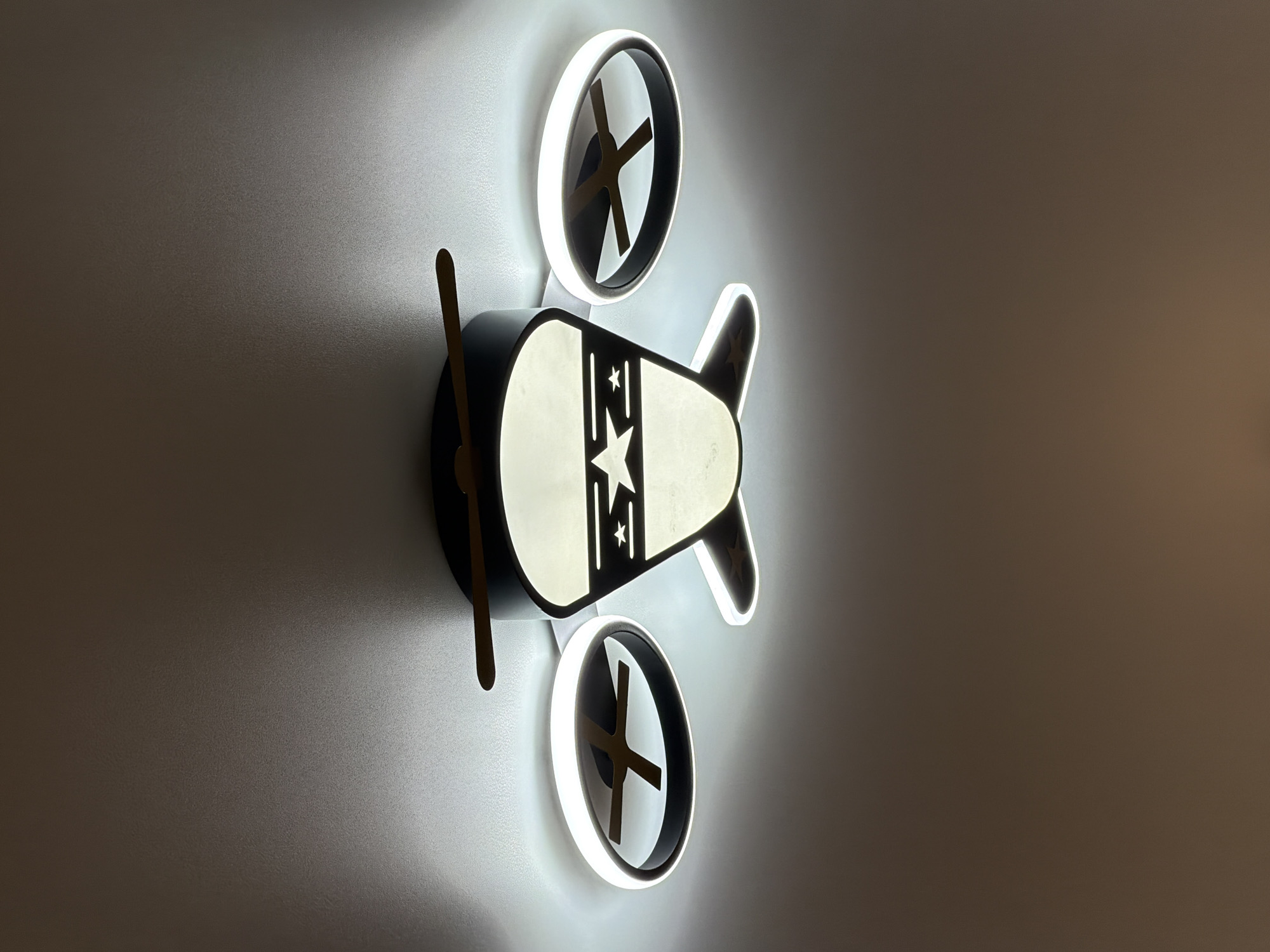 The image shows a ceiling lamp shaped like an airplane. The lamp has a dark central body with a white, illuminated section featuring stars and stripes. It has two circular, illuminated "rotors" or "wings" on either side, each with a dark cross-shaped element inside. Two wooden propeller blades extend from the top of the central body. The lamp is mounted on a white ceiling. The scene is lit by the lamp itself, casting a warm glow downwards and creating shadows on the ceiling. There are no people visible in the image. The location in Chaikovsky, Russia, is not discernible from the image.