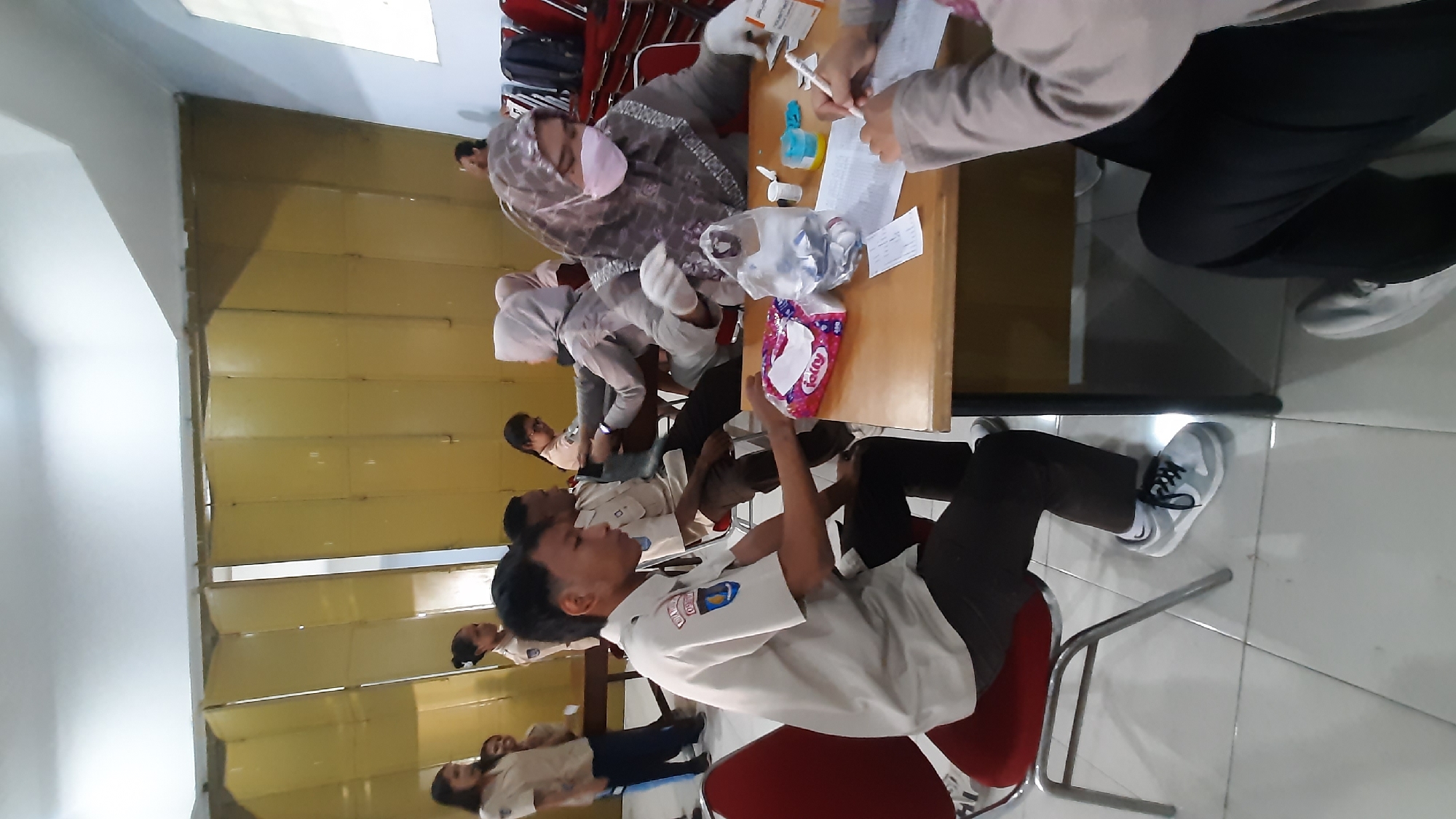 The image depicts an indoor scene, likely a community health event, in Nanggulan, Indonesia. In the foreground, a young man wearing a scout uniform and brown pants sits in a red chair, looking down at a packet of tissues on a wooden table. To his right, a woman wearing a light brown hijab and pink face mask, gloves, and a patterned gray top is seated at the table, appearing to administer a medical procedure to a person whose arm is visible. A second person, also in a scout uniform, is seated behind the woman, partially obscured. In the background, several other individuals, some in similar uniforms, are present, and the environment features a partition wall made of foldable yellow panels. Red chairs are stacked in the background. The floor is tiled with a light gray pattern.