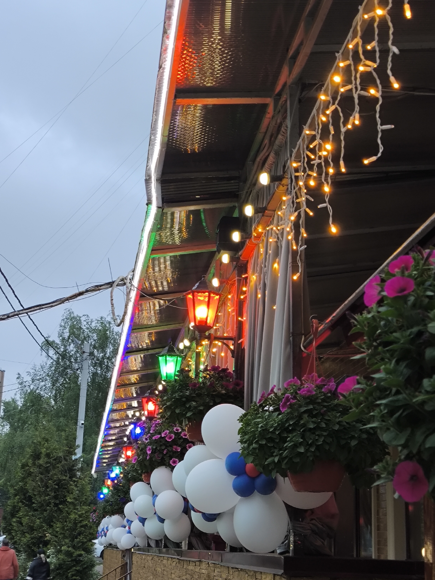 The exterior of a building in Khimki, Russia, is adorned with festive decorations. A string of multicolored lights runs along the roofline, reflecting off the metallic surface beneath. Additional strings of warm-toned lights hang down, casting a glow on the scene. The building is also decorated with a series of lanterns, illuminated in red and green. Several hanging flower pots with pink flowers are visible. White balloons with blue accents are clustered along the ledge of the building. In the background, trees can be seen, with utility lines stretching across the sky. Two people, one wearing an orange jacket and another wearing a black jacket, are partially visible in the lower left corner.