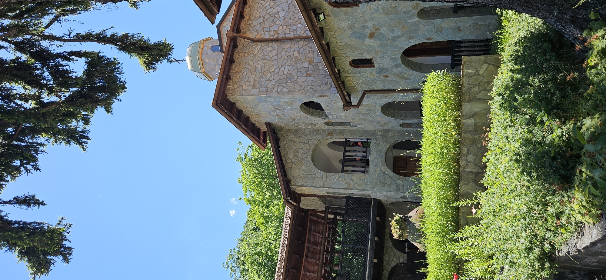 In Ovchartsi, Bulgaria, this outdoor scene features a multi-story stone building with arched windows and doorways. The building has a prominent dome topped with a cross, suggesting it is a religious structure, possibly a church or monastery. Green foliage, including trees and manicured bushes, surrounds the building, with a tree trunk visible on the right. A wooden balcony with a railing is visible on the second story. The sky above is a clear blue.