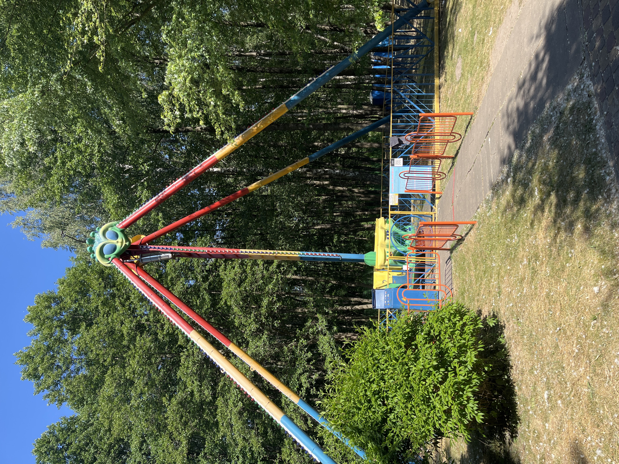 A colorful amusement park ride with a large, green, bug-like head at its apex stands in an outdoor setting surrounded by trees. The ride has four support beams painted red, yellow, and blue, extending outwards from the central structure. A chain of lights hangs from the center of the ride. In the foreground, a paved path leads towards the ride, bordered by a low, orange fence and green bushes. The ground is covered in patchy grass with some dry, light-colored debris scattered across it. Behind the ride, a dense forest of trees forms a backdrop, with a clear blue sky visible above. A blue and yellow fence encloses a portion of the area around the ride's base.