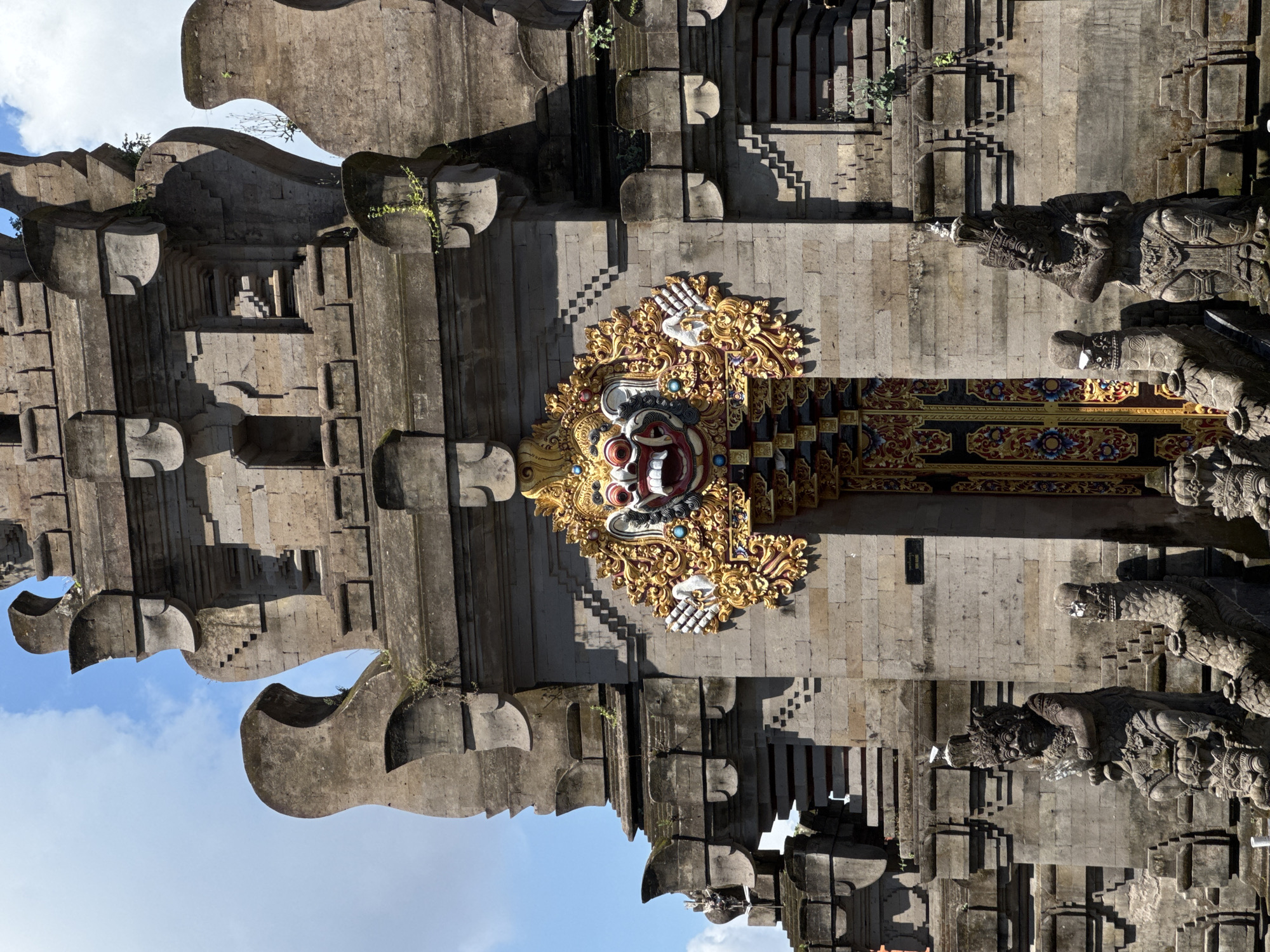 This image depicts a tall, intricately carved stone temple gate or entrance, characteristic of Balinese Hindu architecture, set under a bright blue sky. The structure is built from large, grey stone blocks, forming a majestic, multi-tiered facade.
The central feature is a highly ornate, gilded carving positioned above a dark, richly decorated wooden doorway. This carving presents a stylized mask-like face, possibly a Boma head, with prominent red, white, and black elements, flanked by elaborate gold filigree and details, including stylized white hands.
On either side of the central gateway, massive, stepped stone structures rise, featuring decorative cut-outs and alcoves. At the base of the gate, flanking the entrance, stand imposing stone statues of guardian figures. The one on the right is clearly visible, depicting a powerful mythological being in traditional attire, holding what appears to be a staff or weapon. Behind the main gate, a series of stone steps ascends into the inner temple complex.
Sparse green foliage, likely small vines or moss, grows on parts of the ancient stonework, hinting at its age and natural integration with the surroundings. The scene is illuminated by bright sunlight, casting sharp shadows that highlight the intricate details of the carvings and architectural elements, suggesting it is midday or early afternoon on a clear day. The sky is a vibrant blue with scattered white clouds, visible through openings in the upper structure.
The architecture and carvings are distinct to Balinese religious sites, indicating its location in North Kuta, Indonesia. A small, dark rectangular sign with indistinct white text is visible on the stone wall above the right guardian statue. No specific activity or event is taking place; the image focuses on the grandeur and detail of the architecture.