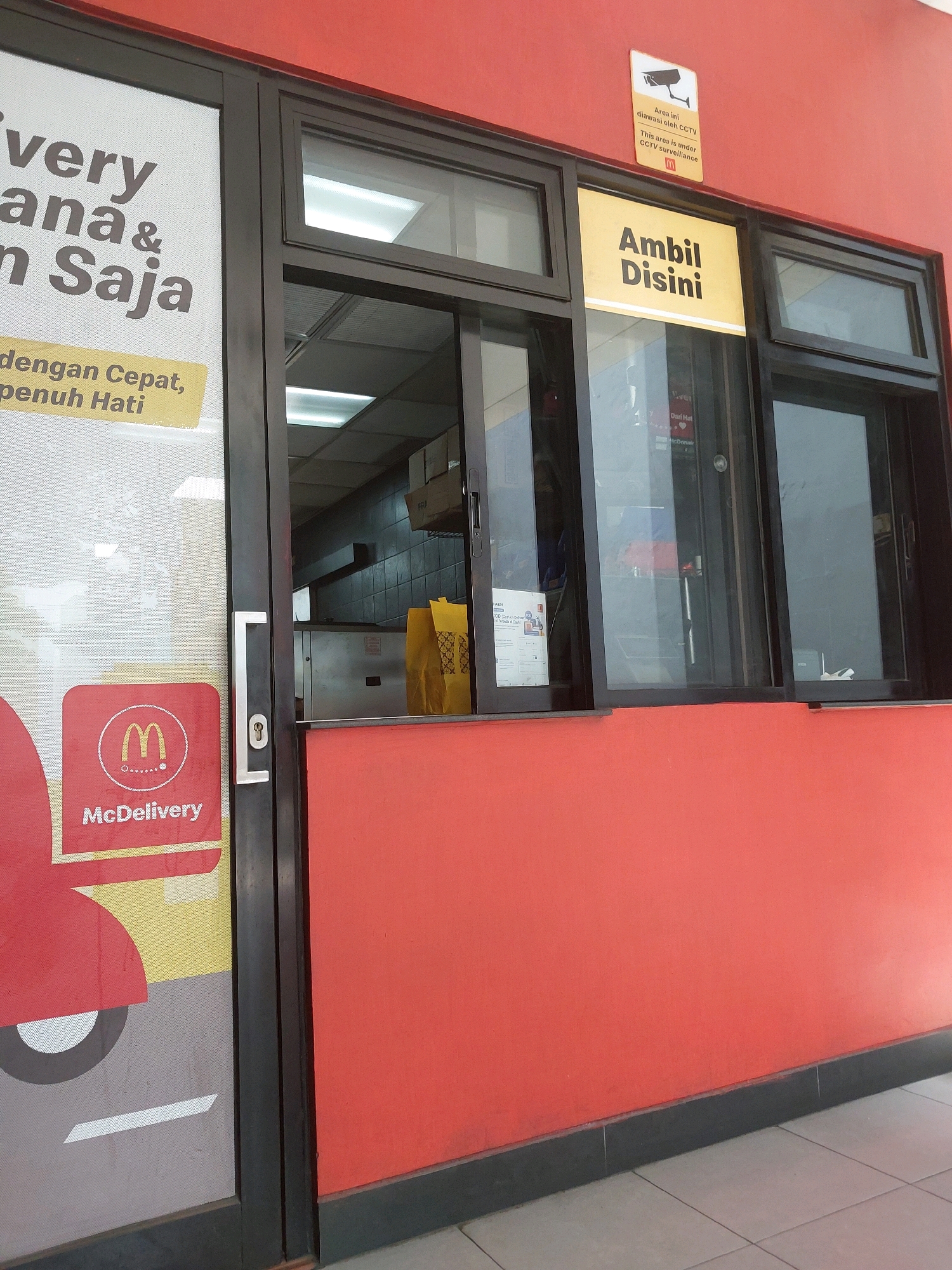 The image captures a close-up view of a McDonald's drive-thru or pick-up window, likely in Malang, Indonesia, given the Indonesian text visible. The building's exterior is painted a vibrant red. A large glass window section is prominent, divided into multiple panes, with some open and others closed. To the left, a glass door with a silver handle and a lock is visible, partially obscuring a large graphic featuring a red vehicle and the "McDelivery" logo, indicating a delivery service.
Above the windows, a yellow sign with bold black text reads "Ambil Disini" which translates to "Pick Up Here." A smaller sign above this one, also with some Indonesian text and English text, states "Area ini diawasi oleh CCTV" and "This area is under CCTV surveillance," accompanied by a CCTV camera icon and the McDonald's logo.
Through the open window, the interior of the McDonald's kitchen can be seen, with stainless steel equipment, tiled walls, and a yellow McDonald's bag on a counter. The floor inside appears to be tiled. The scene is set outdoors, with the red wall forming the main subject, suggesting it's a storefront or service area. The lighting is bright, indicating daytime. No people are clearly visible.