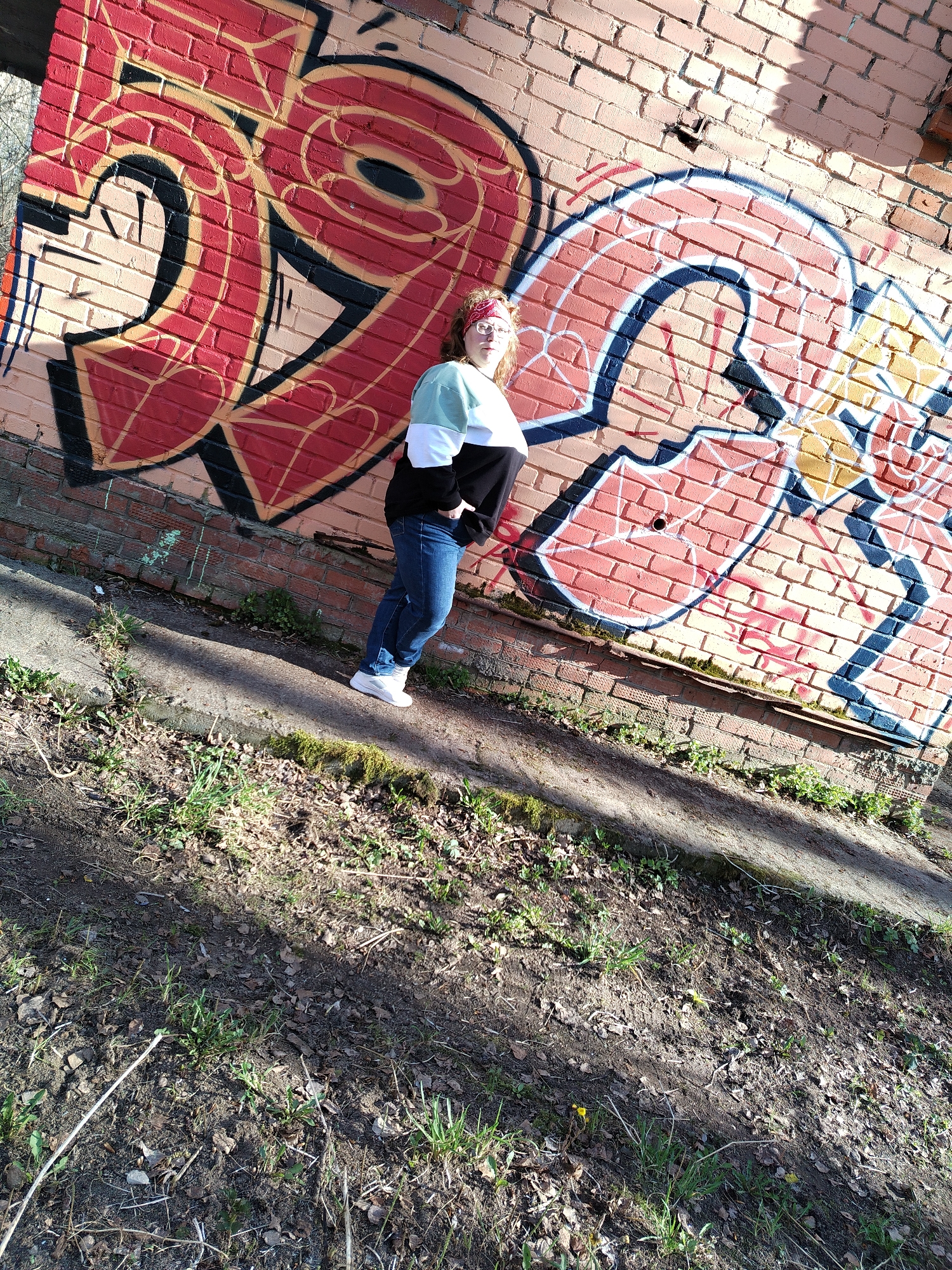 The photograph depicts a young woman standing in front of a brick wall covered in graffiti. The woman is wearing jeans, white sneakers, and a sweater with black and white stripes. She has curly hair and a red headband. The graffiti on the wall is in red, black, and yellow. The ground in front of the wall is covered in grass and dirt. The photograph was taken outdoors in the city of Chaikovsky, Russia.