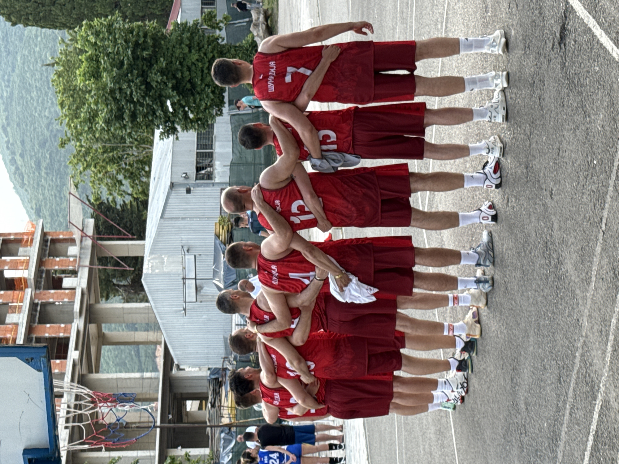 A group of ten young men, appearing to be a basketball team, are standing in a line on an outdoor basketball court, with their arms around each other's shoulders. They are all wearing matching red basketball jerseys and shorts. The jerseys have Cyrillic lettering on the back, with numbers visible on some of them, including 12, 10, 15, and 7. The court is asphalt, with white lines marking the boundaries. In the background, there is an unfinished concrete building under construction, a corrugated metal structure, and lush green trees and hills. The sky is overcast. The scene appears to be in Budva, Montenegro, as indicated by the backdrop of mountainous terrain commonly found in the region.