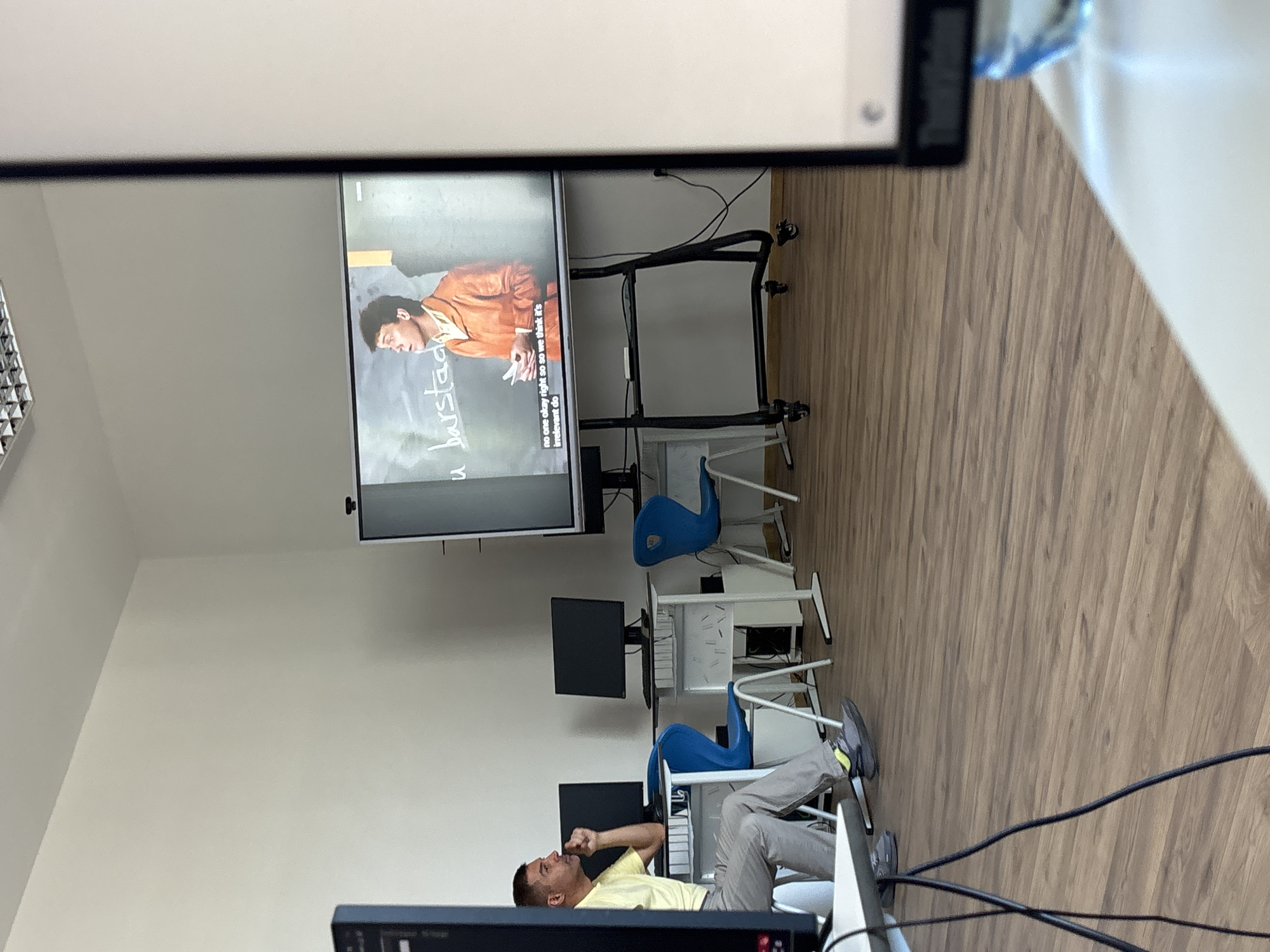 In a classroom setting, a man in a yellow t-shirt and grey pants sits at a desk, looking towards a large screen displaying a video of a man in an orange jacket. The screen shows text that reads "u barstad" and a subtitle that says "no one okay right so so we think it's irrelevant do". The room has white walls, a wooden floor, and several desks with computer monitors and blue chairs. The overall scene suggests a lecture or presentation is taking place. The specific location in Shumen, Bulgaria, cannot be determined from the image alone.