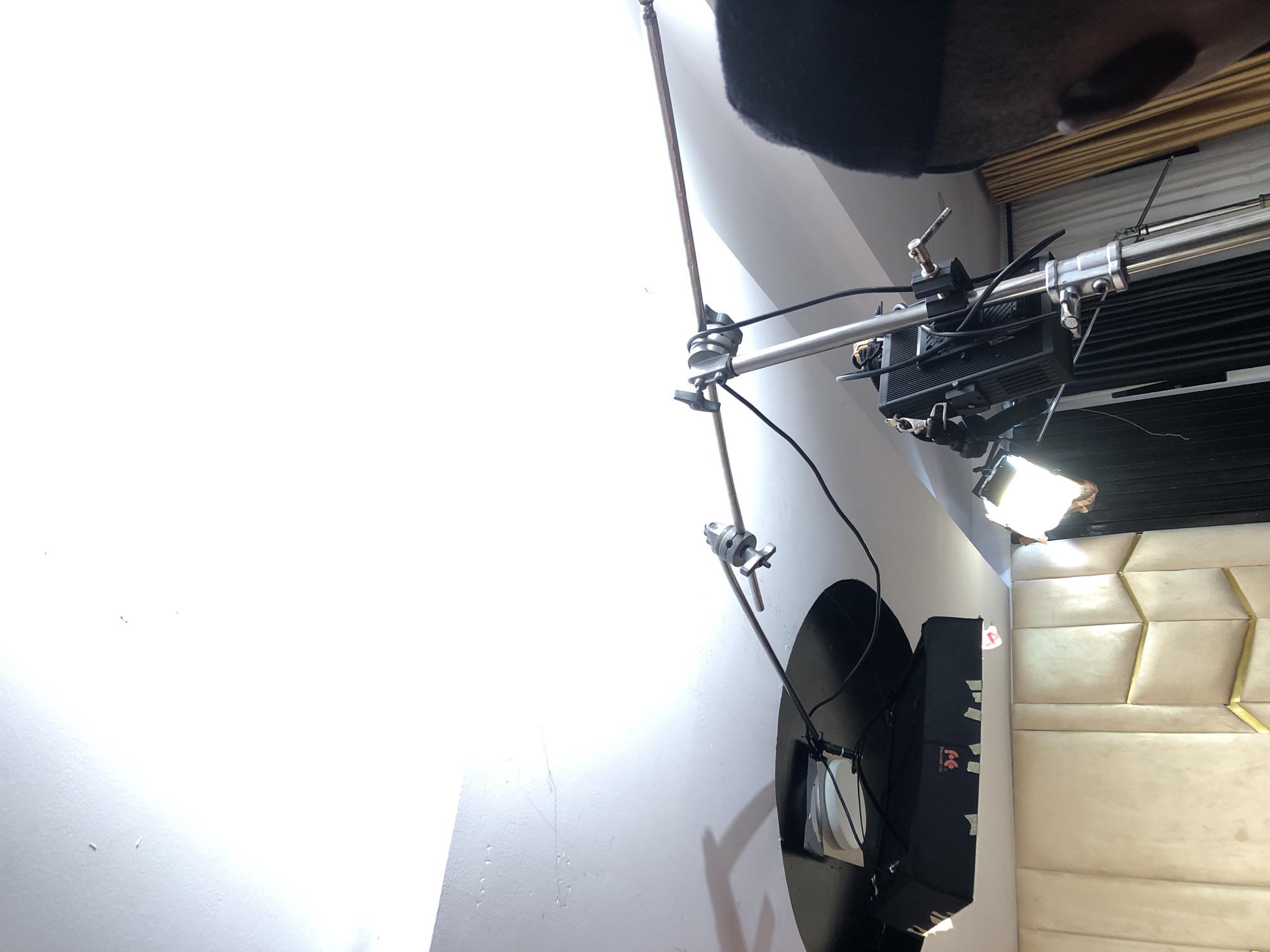 The image displays an indoor setting featuring professional video or photography lighting equipment, suggesting an active shoot or setup. On the left, a bright white wall serves as a backdrop. A heavy-duty light stand, possibly a C-stand, extends a boom arm horizontally from the mid-left towards the center.
Attached to this boom arm is a large, black parabolic softbox, identifiable by the visible text "APUTURE Light Dome II." Below and slightly to its right, a rectangular LED light panel is mounted, emitting bright illumination. The word "feel" is partially visible on this panel, indicating its brand. Further along the boom arm, another professional light fixture, possibly an LED panel with barn doors, is clamped, shining intensely towards the right side of the frame. Various cables, clamps, and rigging components are visible, indicating a complex lighting arrangement.
On the right side of the frame, a luxurious, tufted bed headboard is prominent. It features a light beige or cream color with a distinctive geometric pattern formed by raised panels and golden or brass-colored trim, resembling hexagons or diamonds. Above the headboard, a dark ceiling or overhead structure is visible, with a smaller, rectangular light fixture also directing light downwards. A dark, fuzzy object, likely a person's head or hair, appears at the very top center of the frame, suggesting the presence of an individual near the camera or subject. The scene combines professional studio equipment with domestic furnishing, indicating either a bedroom used as a shooting location or a studio set designed to resemble one.