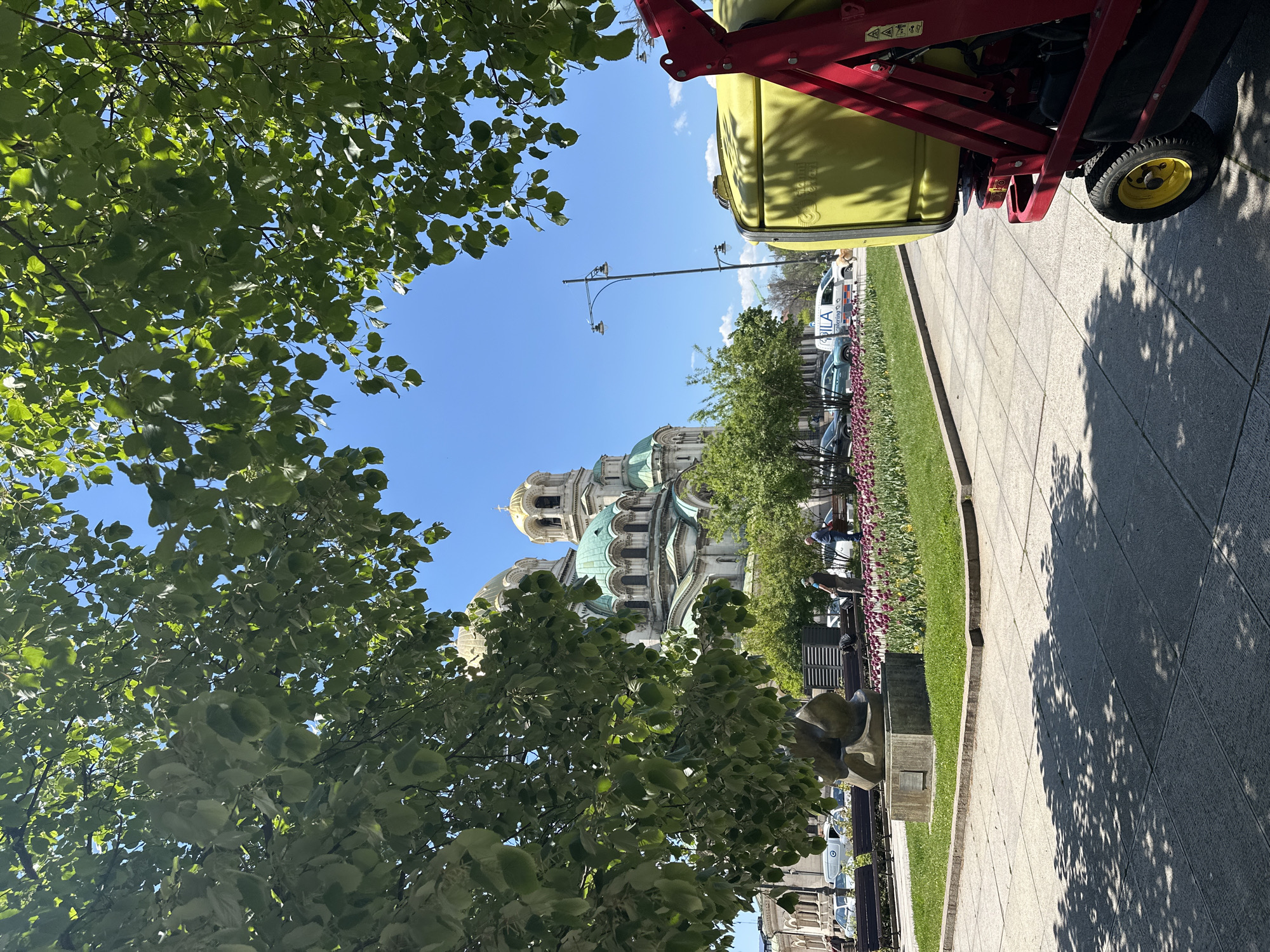 The image shows a partial view of Alexander Nevsky Cathedral in Sofia, Bulgaria, framed by trees on the left and a piece of yellow maintenance equipment on the upper right. The Cathedral's golden domes and green roof are visible in the background. A street lamp is just in front of the building. In the midground, there are flowers, a lawn, a sidewalk, a sculpture, people on benches, and parked cars. The sky is blue and clear. The sidewalk on the right side has shadows from the tree on the left.