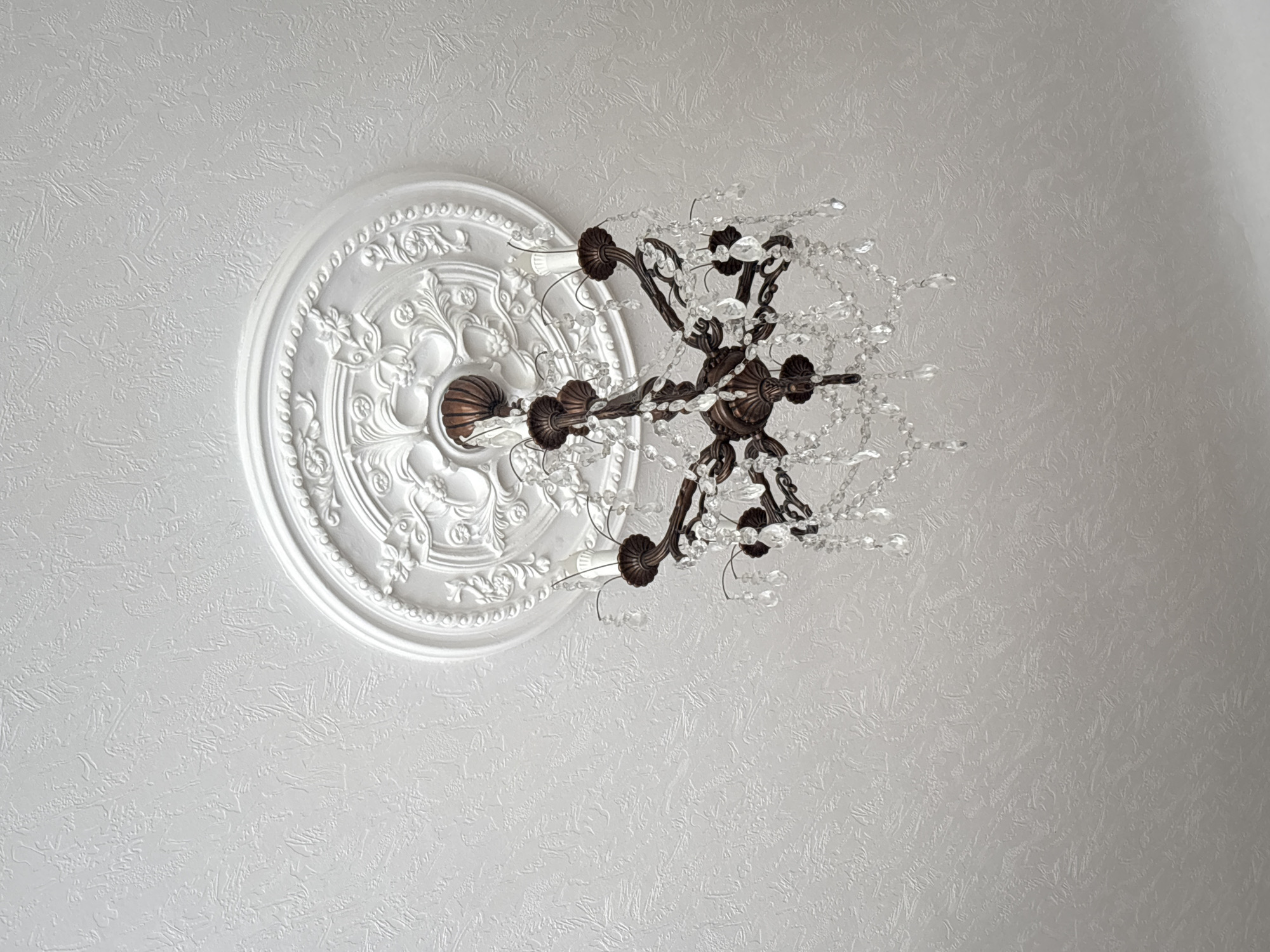 An ornate, dark metal chandelier with crystal accents hangs from a white ceiling with a textured finish. The chandelier has multiple arms holding candle-shaped light fixtures, and is adorned with numerous clear, faceted crystals that reflect light. A decorative white ceiling medallion with intricate relief patterns is visible above the chandelier. The image appears to be taken indoors, focusing on the ceiling and light fixture. There are no people or discernible actions in the image. No information about the city of Sagopshi, Russia, can be inferred from the image.
