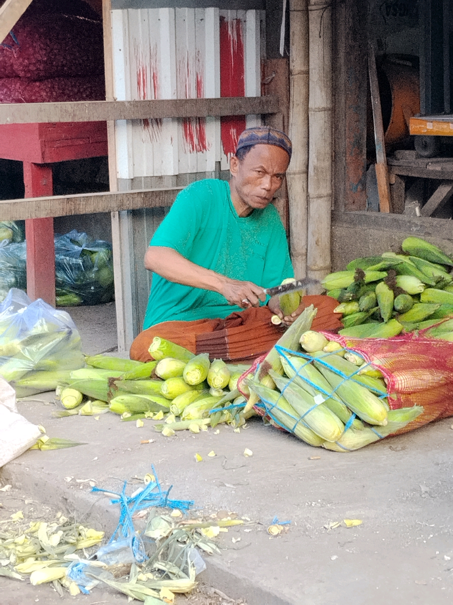 An elderly Indonesian man, wearing a green t-shirt and a dark cap, sits on the ground in front of a stall, preparing corn. He is peeling corn cobs with a knife. A large pile of corn cobs is in front of him, some in a red mesh bag tied with blue string, and others scattered on the ground. Behind him, bags of produce and wooden structures are visible, suggesting an outdoor market setting in Malang, Indonesia. The ground is dirty and littered with husks and plastic debris.