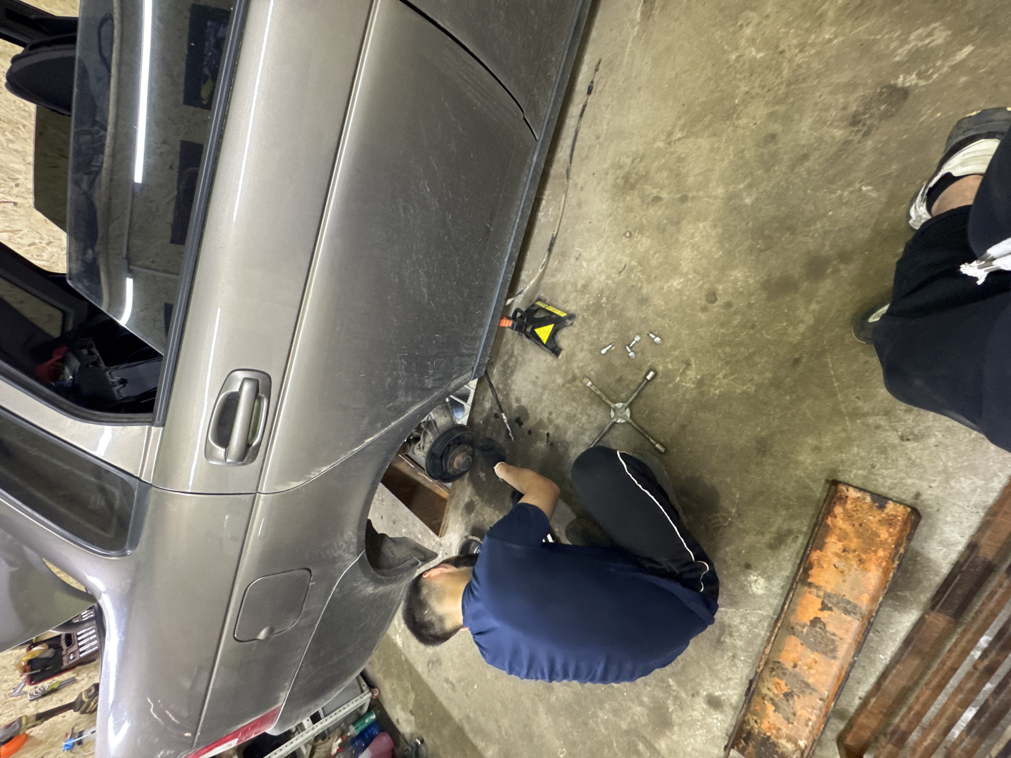 This image depicts an indoor scene in Perm, Russia, specifically within a garage or auto repair shop.
The central subject is a grey/tan metallic sedan, which is currently elevated on its right rear side, indicating a repair or maintenance operation. The rear passenger-side wheel has been removed, exposing the brake drum and hub assembly. A yellow and black jack stand provides primary support under the car's frame, while a wooden plank offers additional stabilization under the suspension component. The rear passenger door is ajar, revealing a dark interior. The car's body panels, particularly the lower sections, are visibly dusty or dirty. In the upper left background, the car's trunk lid appears to be open or significantly elevated.
A male individual, presumed to be a mechanic, is crouched on the concrete floor facing away from the camera, positioned directly next to the exposed wheel well. He has short dark hair and is wearing a dark blue short-sleeved t-shirt, dark athletic pants with a white stripe down the outer seam, and dark work gloves. His hands are actively engaged with the brake components, suggesting he is performing a repair.
On the floor near the worker, a cross-shaped lug wrench and several silver lug nuts/bolts are scattered. A dark cable runs along the ground beneath the car, extending towards the jack stand.
In the lower right corner, the lower legs and feet of a second person are visible. This individual is wearing black pants and light-colored sneakers with black accents, suggesting they are seated or standing very low to the ground, observing the work.
The environment features a stained, uneven concrete floor. The background includes a light-colored wall, possibly wood paneling, on which various hand tools, including wrenches and sockets, are neatly hung. A red tool bag or case is also visible on this wall. In the far left background, a rack holds multiple colorful plastic bottles, likely containing various automotive fluids or cleaning supplies. Two long, rectangular, heavily rusted metal objects lie on the floor in the lower left foreground. Fluorescent lights are reflected in the car's passenger window.
