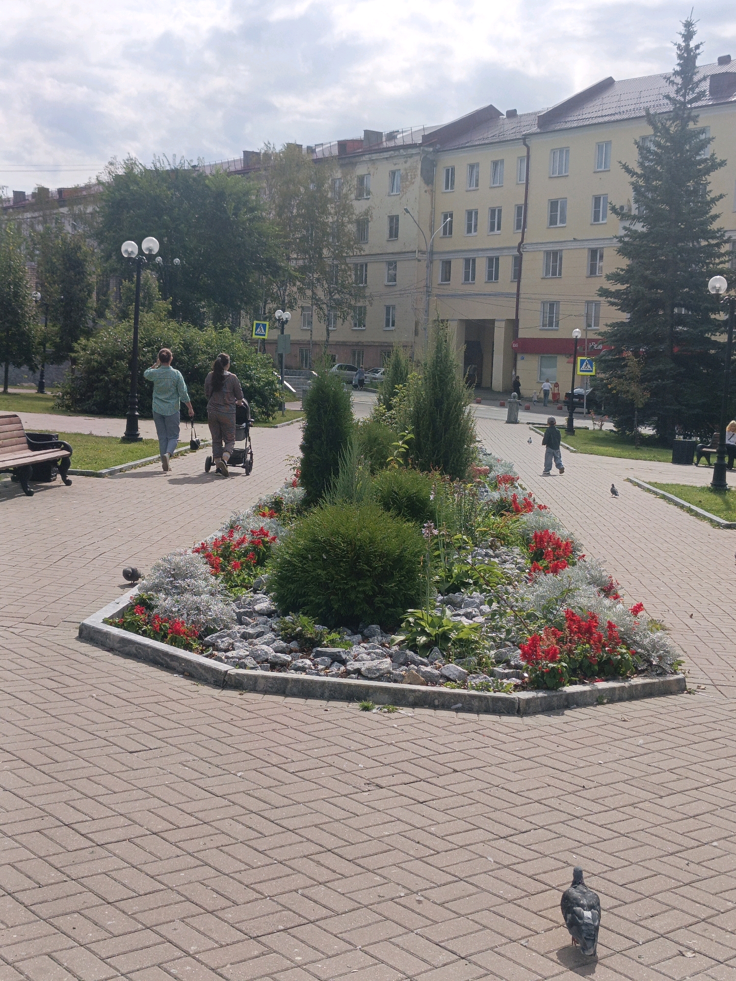 The image depicts a public park area with a paved walkway in the foreground. The walkway is made of reddish-brown interlocking bricks laid in a herringbone pattern. A grey pigeon is seen walking on the bricks in the lower right quadrant. To the left of the walkway, a flower bed is visible, bordered by concrete and filled with grey stones, green foliage, and numerous small red flowers. A larger, rounded green bush is centrally located in the flower bed. In the middle ground, two women are walking away from the viewer on the paved path. One woman wears a green and white plaid shirt and grey pants, while the other wears a patterned jumpsuit and pushes a stroller. Behind them, several trees and lampposts are present. In the background, a light yellow, multi-story building with a red-tiled roof stands prominently. The sky is partly cloudy. This scene is consistent with a public urban space in Satka, Russia, likely a park or pedestrian zone.