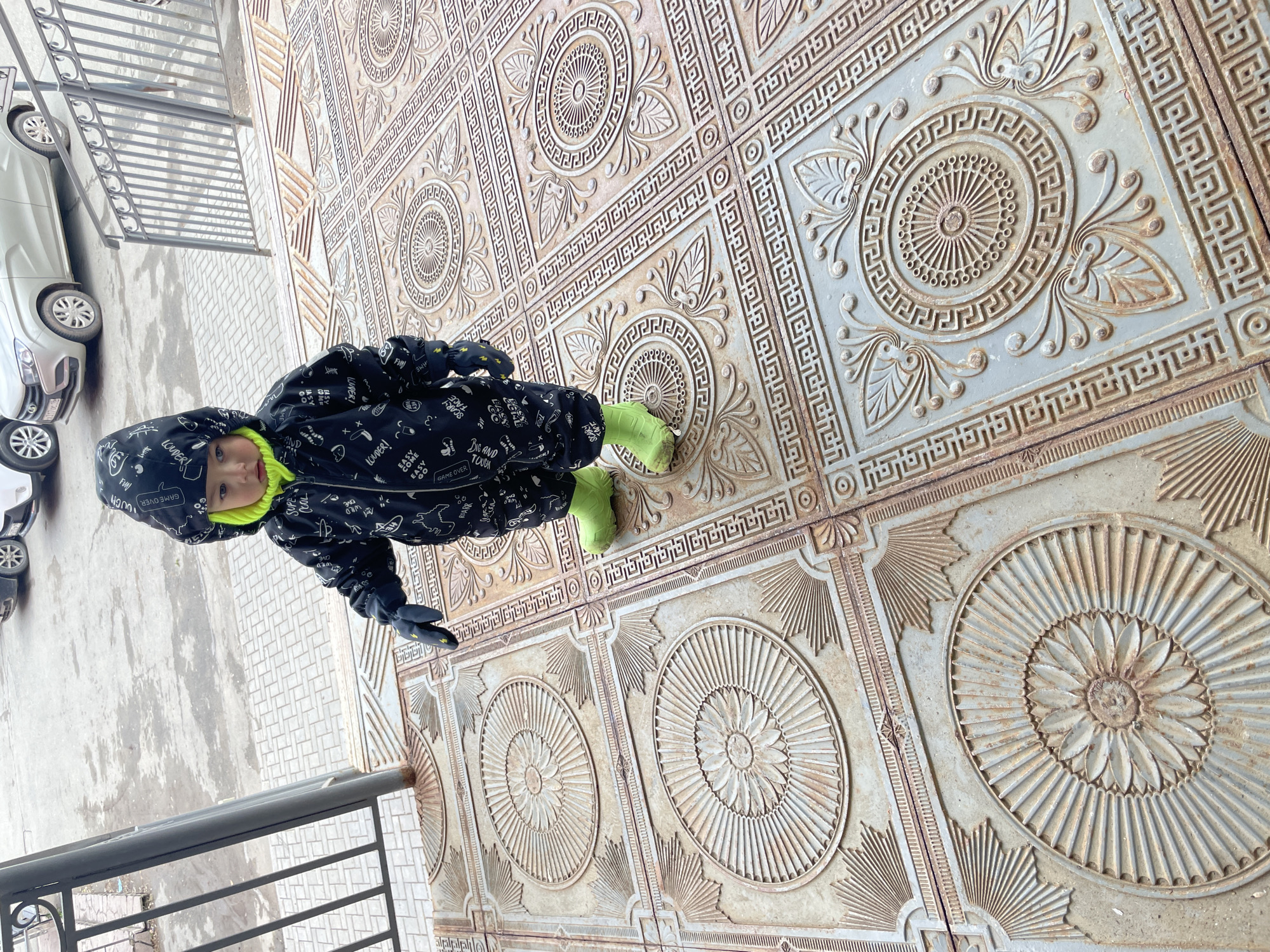 A young child stands on a highly decorative, patterned outdoor surface in Izhevsk, Russia, captured from an overhead perspective. The child, likely a toddler, is dressed for cold weather in a dark-colored, hooded snowsuit featuring various words and phrases such as "WILD," "COOL," "GAME OVER," "LOUDER!", and "TOUGH." They wear a bright lime green or yellow high-neck top beneath the suit and matching lime green boots, along with dark gloves.
The unique ground beneath the child consists of large, intricately patterned square tiles, possibly made of cast iron or concrete. Each tile typically features geometric borders, including Greek key patterns, surrounding a central circular medallion with radial designs, resembling stylized sunbursts or flowers. The surface shows signs of age and weathering, with some discoloration or rust.
To the side, a silver car is partially visible, parked on a light-colored paved area with a cobblestone-like pattern. A dark metal railing separates the decorative ground from the paved section, suggesting an outdoor space such as a balcony, terrace, or elevated walkway overlooking a street or parking area. The soft, diffused lighting indicates an overcast day, consistent with the child's warm attire, implying a cold season like winter or early spring/late autumn. The child looks directly towards the camera with a neutral expression.