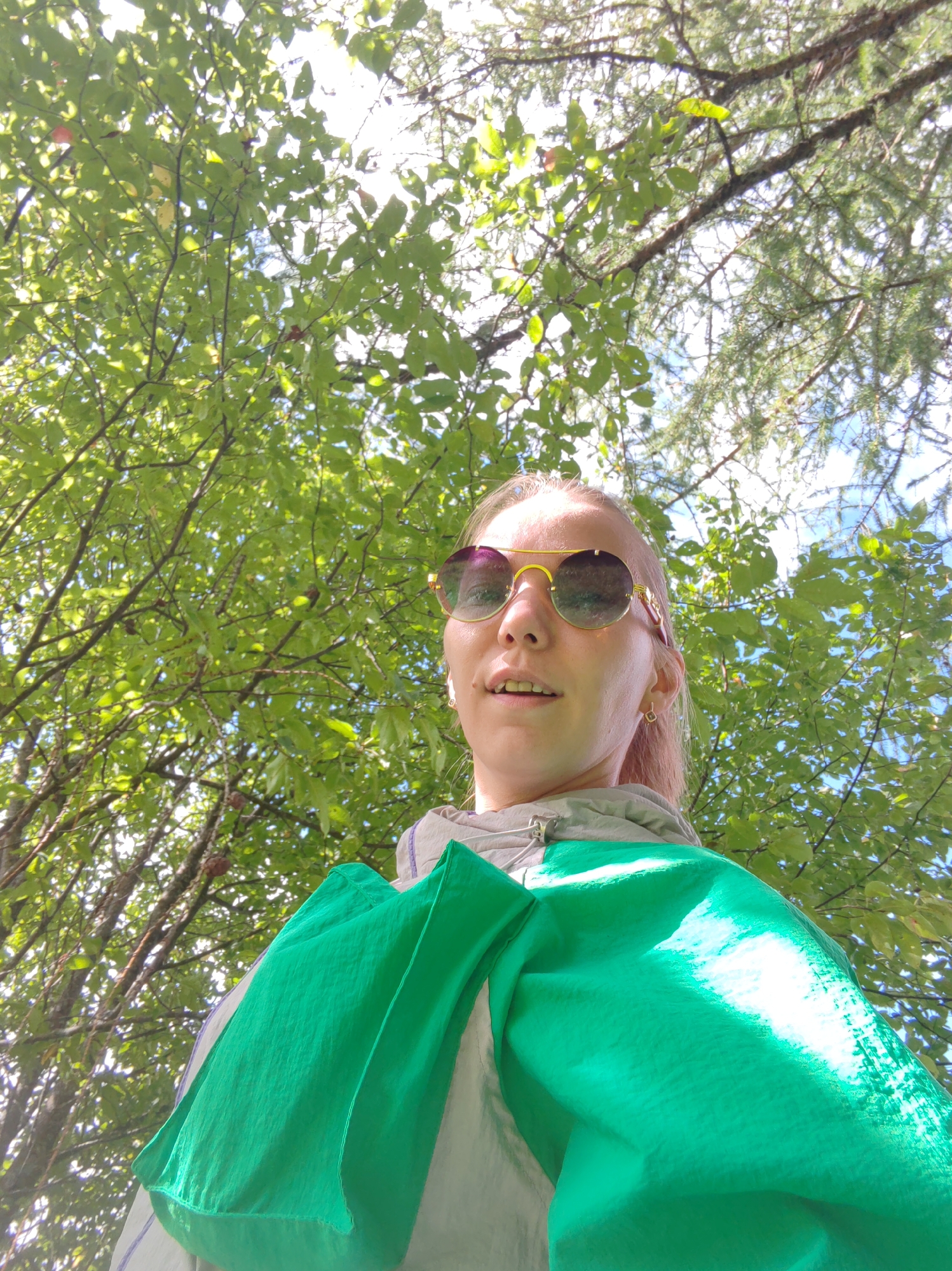 A young woman, with blonde hair and pink undertones, is captured from a low angle, looking up at the camera. She is wearing sunglasses with gold frames and a gray hooded jacket. A bright green fabric is draped over her shoulders and chest. The background is dominated by lush green foliage and branches of trees, with dappled sunlight filtering through, suggesting an outdoor setting. The sky is visible in patches between the leaves. The scene appears to be in a natural environment, possibly a park or wooded area, and it is not possible to determine the specific location within Чухлэм, Russia, from the visual information provided.