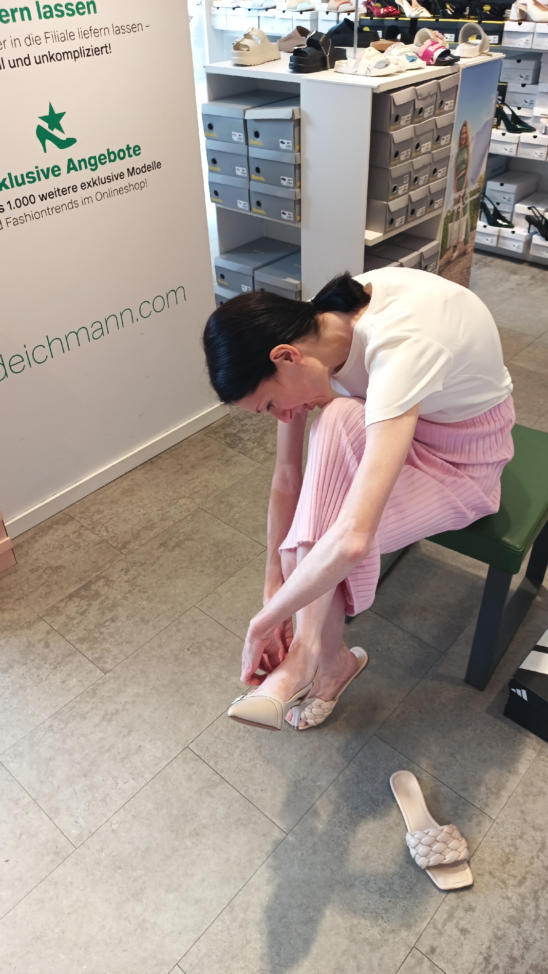 A woman with dark hair tied back in a ponytail, wearing a white t-shirt and a pink pleated skirt, is sitting on a dark green stool in a shoe store. She is hunched over, focusing on her feet as she tries on a pair of light beige, pointed-toe mules with a braided strap. One shoe is already on her left foot, and she is adjusting it with her hands. Another identical shoe lies on the light gray tiled floor next to her right foot. In the background, there are white shelves stocked with shoeboxes and several pairs of shoes displayed on top. To the left of the woman is a white promotional banner with green text and a green leaf logo. Part of the banner reads "exklusive Angebote" and "deichmann.com". The floor is tiled with large, light gray rectangular tiles. The scene appears to be a Deichmann shoe store, a common retail chain, and while the specific location within Nuremberg, Germany, is not explicitly stated, the presence of the Deichmann brand and the typical store environment suggests this context.