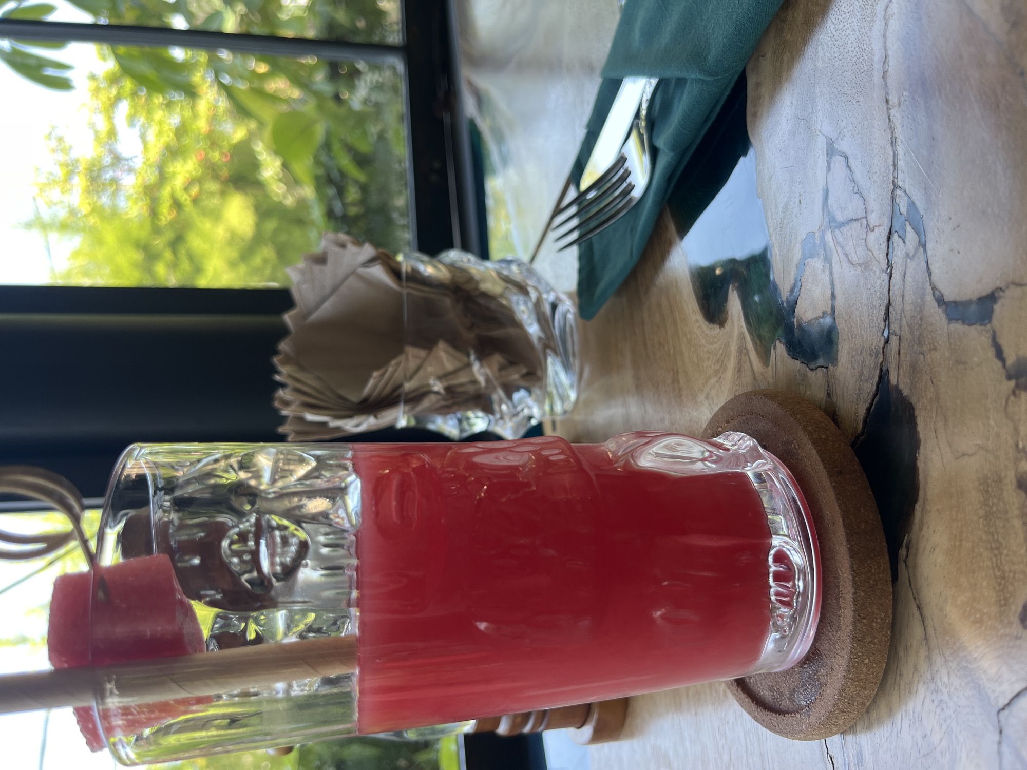 A close-up, medium-shot, eye-level view depicts a glass of watermelon juice, garnished with cubed watermelon on a wooden skewer. The drink sits on a round, cork coaster. In the background, out-of-focus, are a glass container with napkins, silverware wrapped in a green cloth napkin, and a window with green foliage visible outside. The table appears to be made of light-colored wood. The scene appears to be indoors. It is presumed this is a restaurant, located in North Kuta, Indonesia.