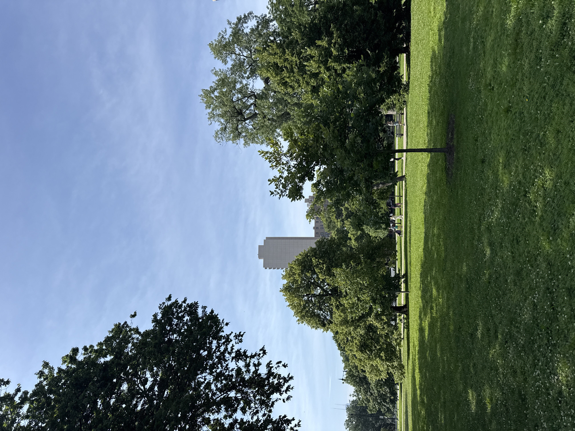 The image depicts a sunlit park with lush green grass and mature trees. A tall, light-colored building rises in the background, partially obscured by the foliage. Several people are scattered across the park, some appearing to sit on benches or walk along a path. The sun casts long shadows, suggesting it is either morning or late afternoon. The sky is a clear blue with a few wispy clouds. The scene conveys a sense of peaceful leisure in an urban green space.