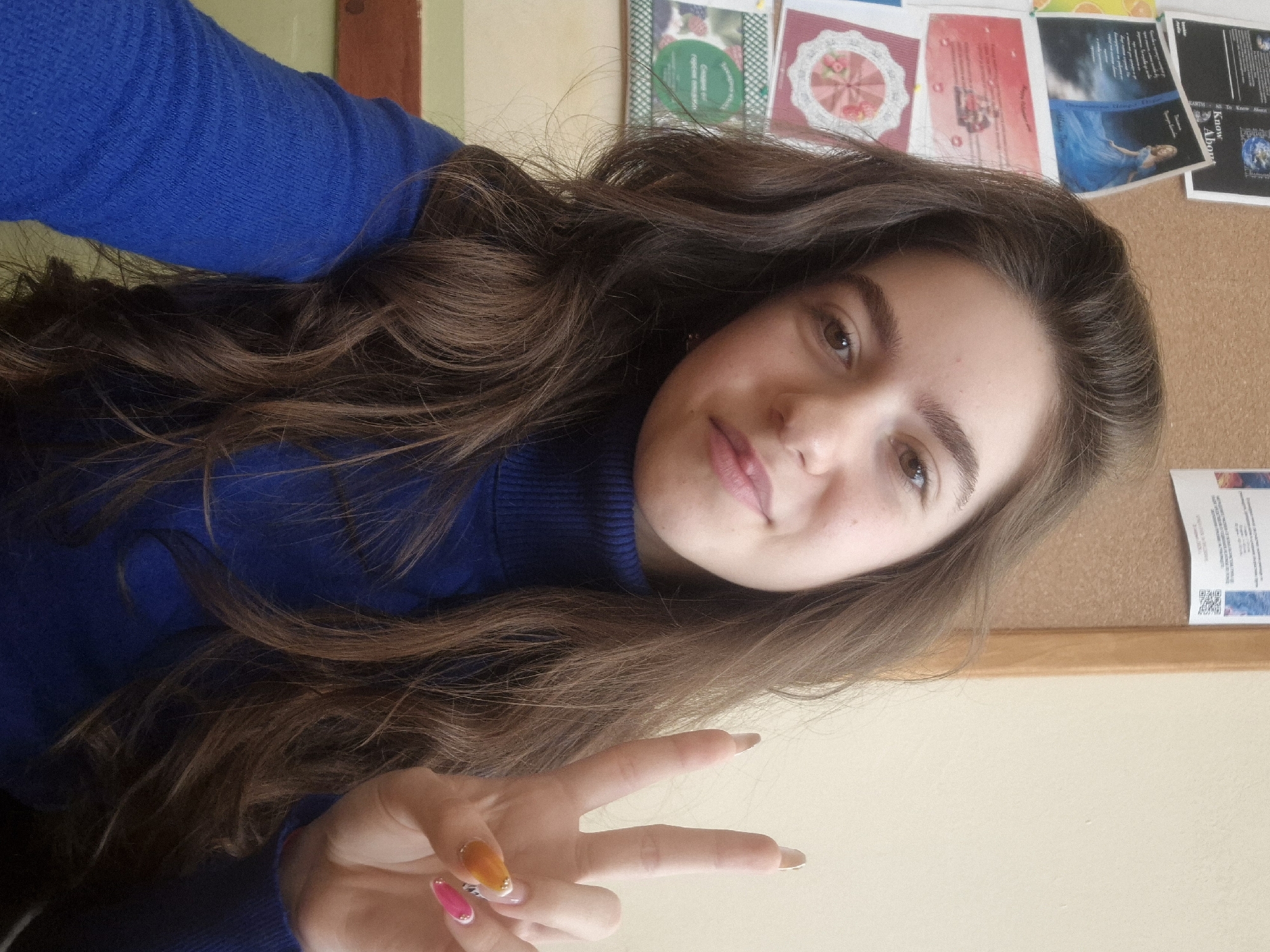 A young woman with long, wavy brown hair is positioned in a selfie-style photograph. She is wearing a blue turtleneck sweater and making a peace sign with her left hand. Her fingernails are painted in different colors. The background includes a corkboard with various papers and cards pinned to it, as well as a portion of a wall and doorframe. The location is Pavlikeni, Bulgaria.