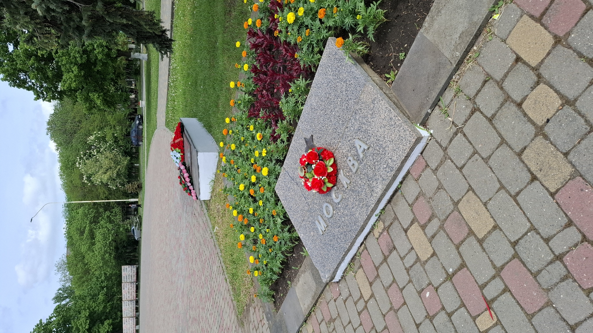 The image shows a memorial site in an urban environment. The memorial features a grey stone slab with the word "MOCKBA" inscribed on it, surrounded by orange and yellow flowers, and a wreath. The memorial is situated next to a brick-paved walkway. In the background, there are trees, grass, and another memorial with flower arrangements. A lamppost and vehicles are also visible in the distance. The sky is overcast.