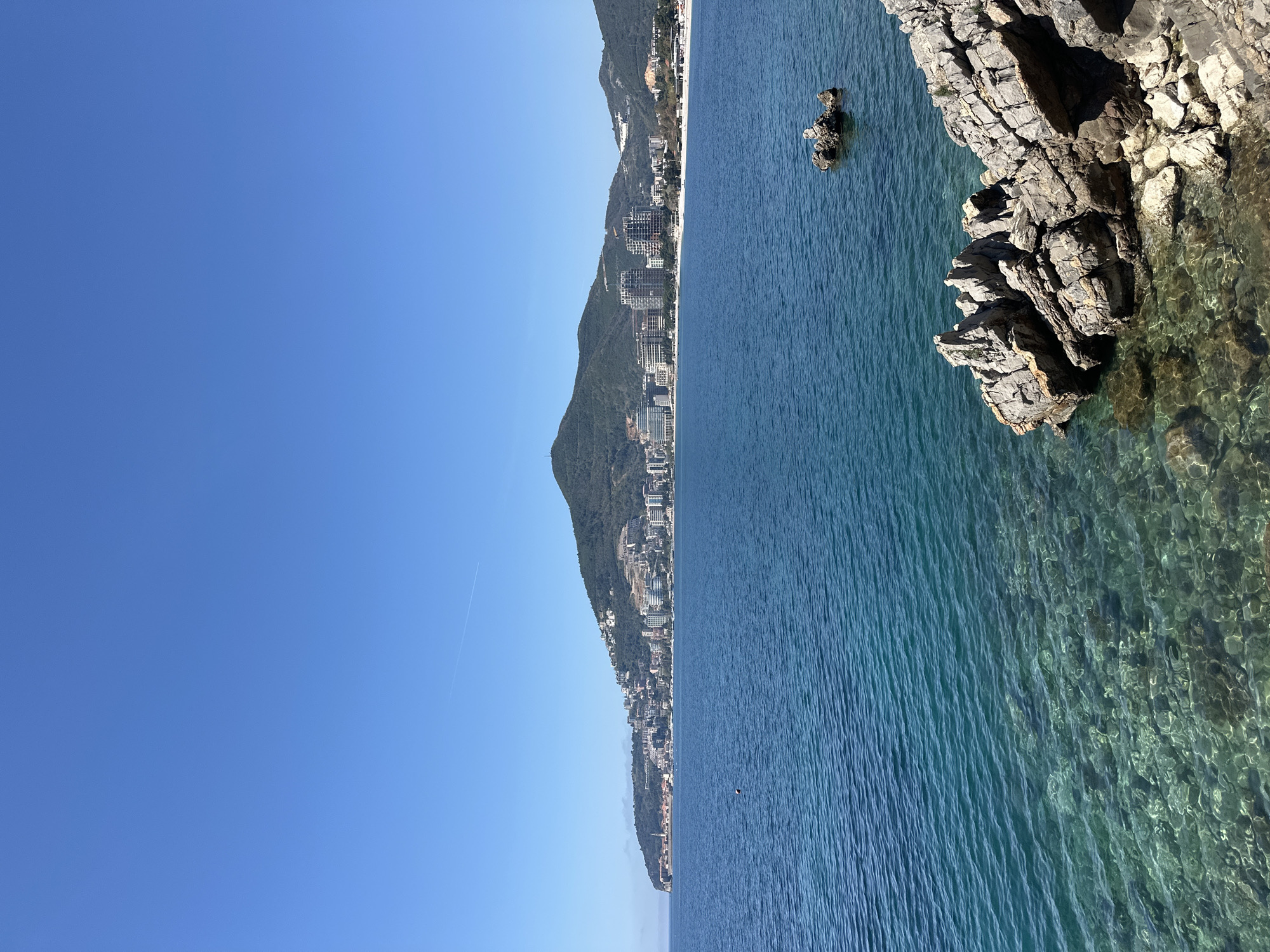 The image depicts a coastal scene in Budva, Montenegro, featuring clear blue waters, rocky cliffs, and a coastal cityscape. The right side of the image showcases rocky formations extending into the water, with visible underwater details due to the water's clarity. Further out, there's a small rock island. Along the coastline to the left, a line of buildings extends along the foot of a hill, suggesting urban development. The sky is clear and blue, occupying a significant portion of the image.