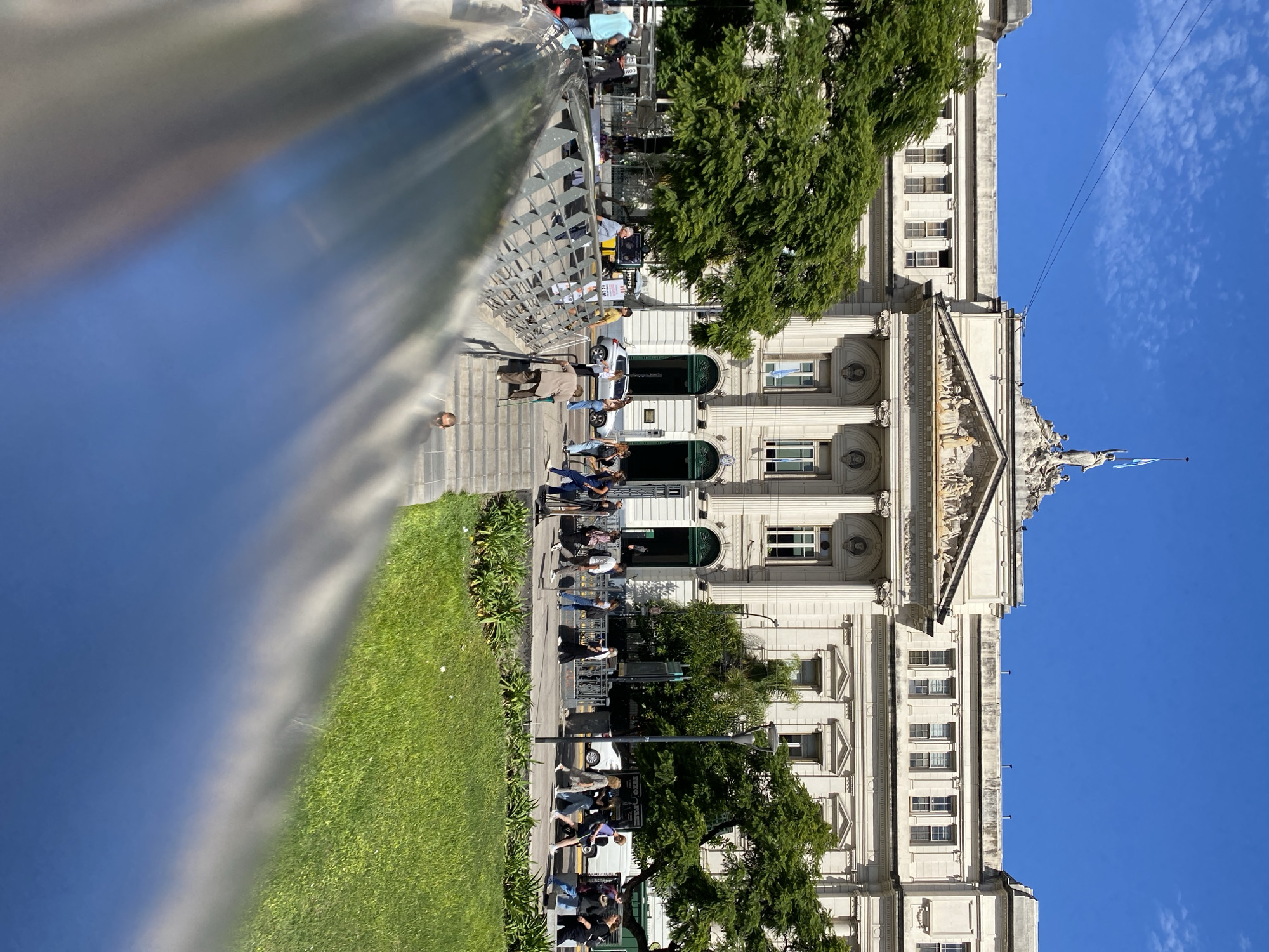 An outdoor, daytime image captures the Faculty of Economic Sciences building of the University of Buenos Aires, located on Avenida Córdoba 2122 in the Recoleta neighborhood of Buenos Aires, Argentina. The prominent neoclassical-style building, constructed of light-colored stone or stucco, dominates the background under a clear blue sky with minimal cirrus clouds. Its central facade features four large columns, an ornate pediment with sculpted figures, and a rooftop statue of a female figure holding a staff, with an Argentine flag flying behind it. The main entrance consists of green double doors.
In the midground, a paved pedestrian area separates the building from a grassy lawn and various trees with full green foliage. Approximately 20-30 individuals are observed walking along the pavement or standing. One individual, dressed in a light-colored top and dark pants, is ascending a set of stone steps on the right, utilizing two walking sticks. Other pedestrians are dressed in casual attire. To the left of the building's entrance, a small market stall with a blue canopy is visible, with a few people standing nearby. A white compact car is parked or slowly moving on a street further back, behind the pedestrians. Black metal railings line portions of the pedestrian area.
The immediate foreground is partially obscured by a highly reflective, curved, metallic surface, possibly a barrier or architectural element, which reflects parts of the sky and a partial, distorted image of a person at the bottom of the steps.