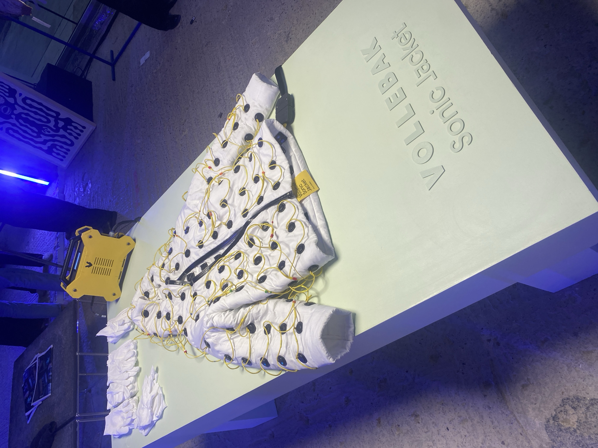 The image shows a "Vollebak Sonic Jacket" on display. The jacket is white with black circular elements connected by yellow wires. It is placed on a light green table with the jacket's name embossed on the surface. A yellow device sits to the left of the jacket, possibly related to its function. White fabric pieces also lie near the jacket. The environment appears to be an indoor exhibition space. There are no visible people in direct interaction with the jacket in the frame, although limbs are visible in the background. Based on the jacket being by Vollebak, and their location, this image is likely to have been taken in London, United Kingdom.
