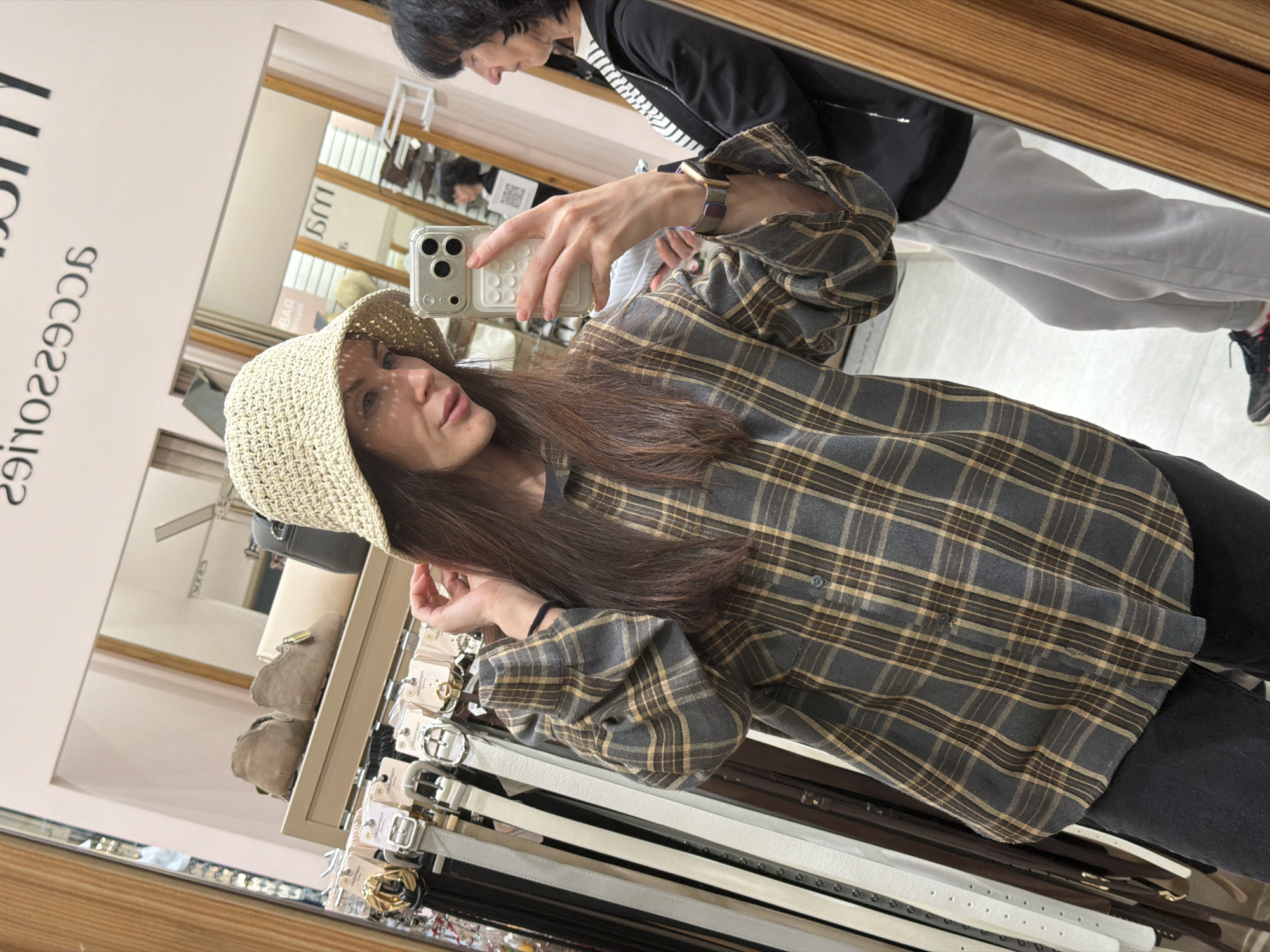 A woman in a straw hat and plaid shirt takes a mirror selfie in a store that appears to sell accessories, possibly in Perm, Russia, based on the store's name and items visible in the reflection. The mirror also reflects another woman in a black jacket and gray pants. The woman is holding a cell phone. Belts and shoes are visible on shelves below the mirror. The overall lighting appears to be artificial.