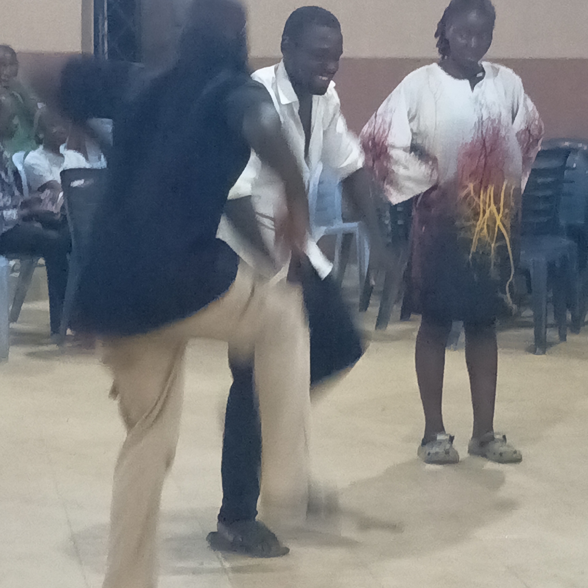 The image depicts three people indoors in what appears to be a hall in Akwanga, Nigeria. A person in a black shirt and tan pants is in the foreground, partially obscuring the view of the other two. The person in the middle is wearing a white shirt, black pants and is smiling. To the right, a woman is wearing a white and red top with a dark skirt that has yellow designs. She is also wearing grey Crocs. Several chairs are in the background, some occupied by other individuals who are partially visible. The floor is a light color. The overall scene has a slightly blurred quality, suggesting movement.