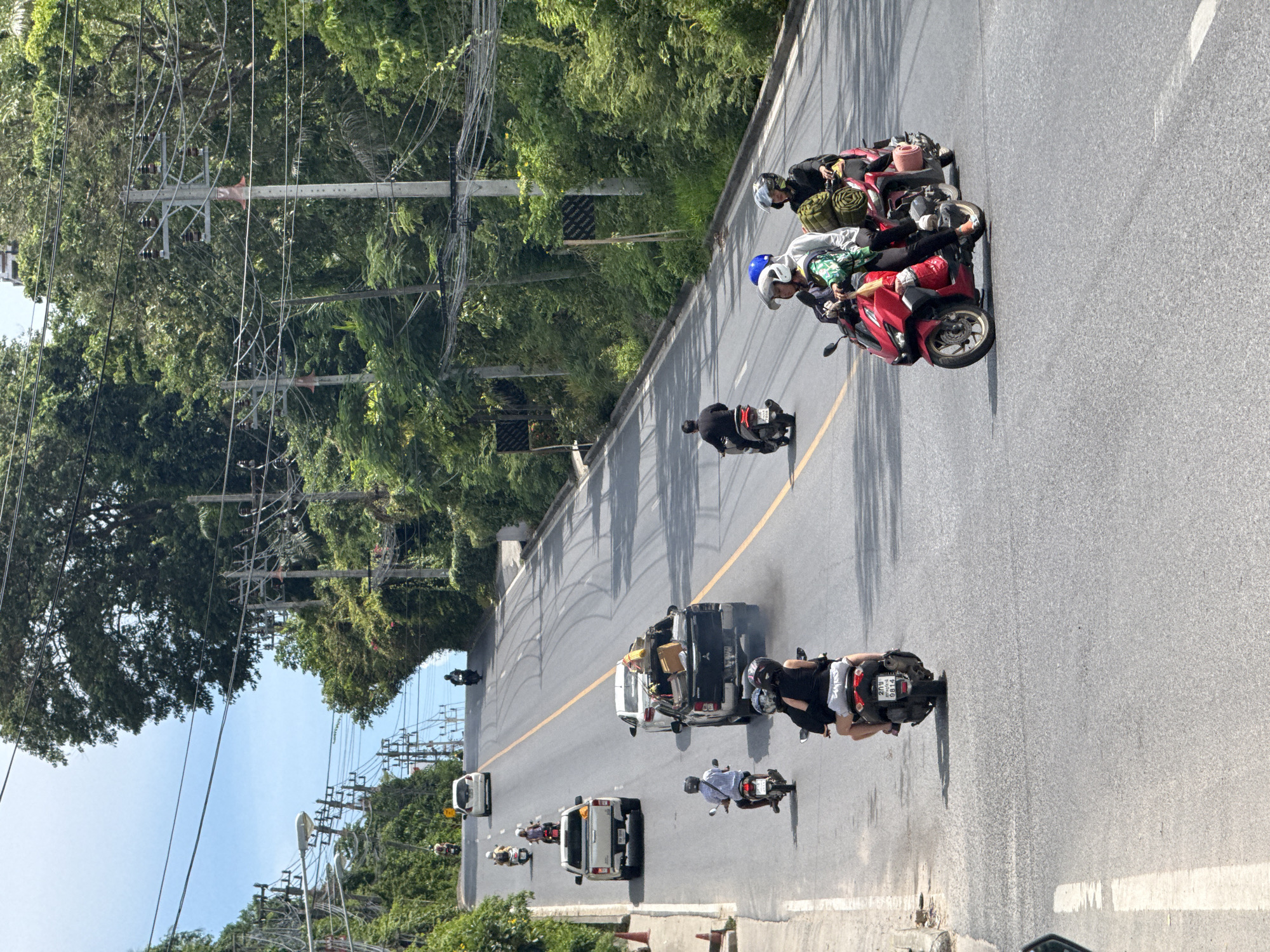 In Baan Chaweng Noi, Thailand, a busy road winds uphill through lush, tropical foliage. The asphalt is marked with a yellow center line and white lane dividers. Numerous motorbikes, a pickup truck, and a car navigate the road. On one motorbike, a woman in a black top and white shorts rides as a passenger, her legs extended forward. The driver, wearing a black helmet, is obscured. Another red motorbike carries three people: a driver in a blue helmet, a passenger in a green and white patterned shirt and white helmet, and another passenger behind them in a dark helmet. A black pickup truck in front of them emits a plume of dark exhaust. Further up the road, another white pickup truck is parked. Power lines crisscross above the roadway, supported by concrete poles. The sky is clear and blue.