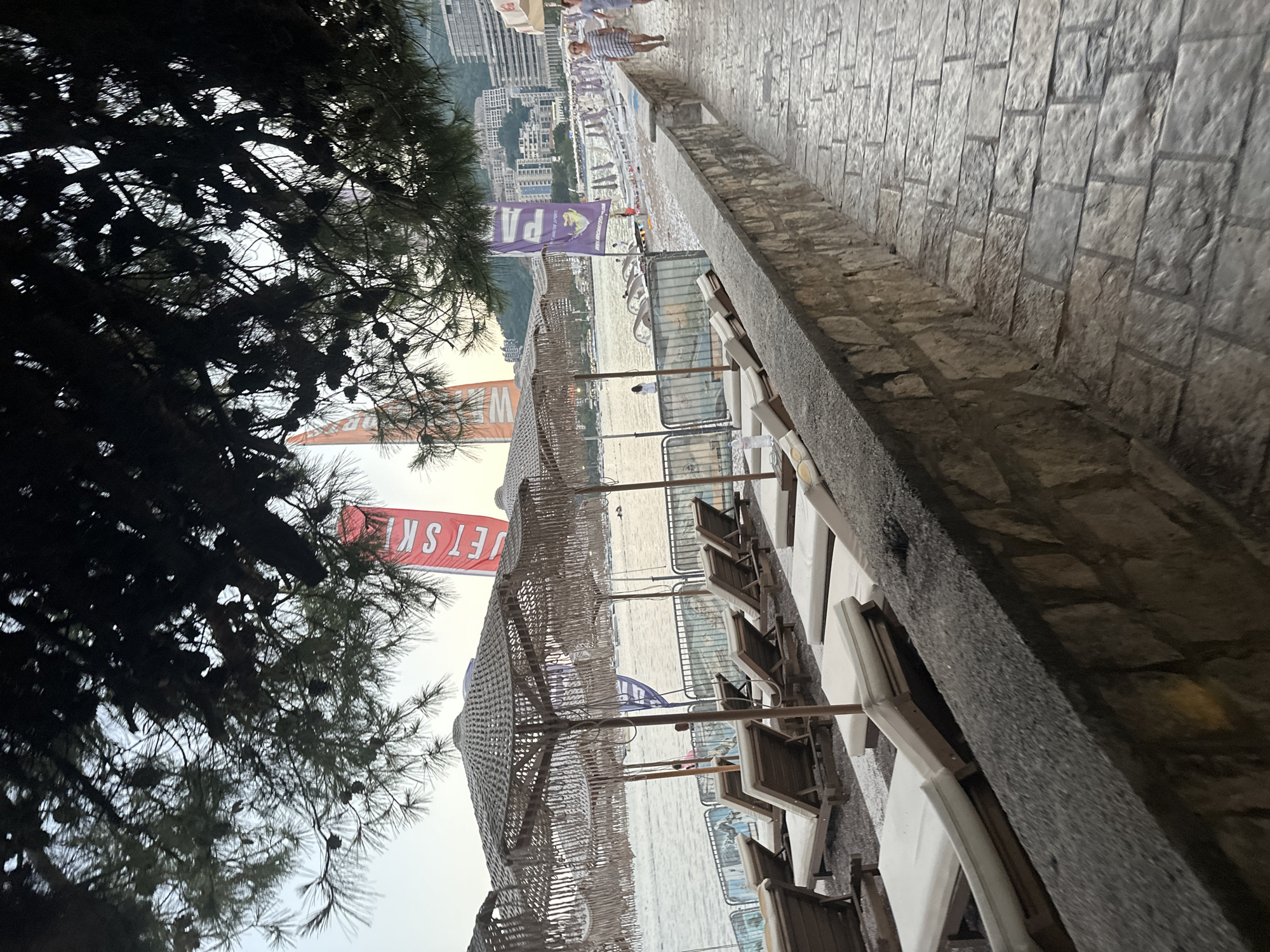 The image depicts a section of the promenade adjacent to a beach in Budva, Montenegro. The foreground features a stone pathway constructed from light-colored rectangular paving stones. To the left of the pathway, a low wall made of rough-hewn stones separates it from a beach area. This beach area is lined with numerous empty lounge chairs and straw-thatched umbrellas, suggesting it is a public beach with amenities. In the background, the calm sea is visible, with a few boats and swimmers in the distance. The cityscape of Budva rises behind the beach, characterized by numerous modern buildings. Two vertical banners, one red and one purple, are prominent on the promenade, with visible text that appears to be advertisements for recreational activities like jet skiing. The overall lighting suggests either early morning or late evening, with soft, diffused light. A couple walks away from the viewer on the right side of the promenade. A few people are scattered on the beach and in the water. The scene conveys a tranquil atmosphere, characteristic of a seaside resort town.