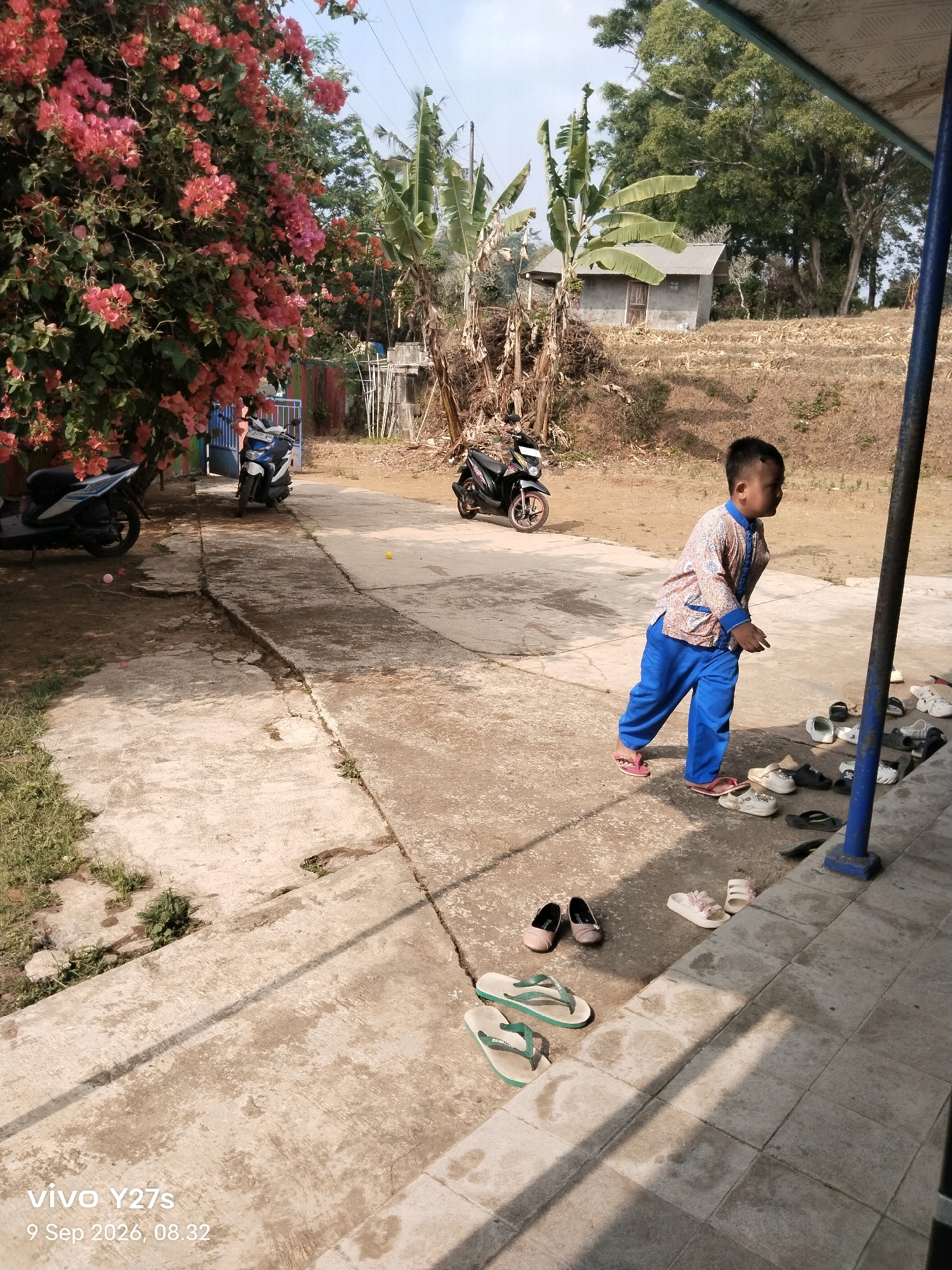 The image depicts an outdoor scene in Tamansari, Indonesia, on September 9, 2026, at 08:32. A young boy, wearing blue pants and a patterned shirt, is walking on a concrete pathway. He is surrounded by various footwear, including flip-flops and sandals, neatly arranged along the edge of the pathway. In the background, a small concrete building with a corrugated metal roof is visible, situated on a terraced hillside covered with dry vegetation. Several motorbikes are parked nearby, and a vibrant bougainvillea bush with pink flowers is in bloom on the left side of the frame. The lighting suggests it is daytime with bright sunlight casting shadows.