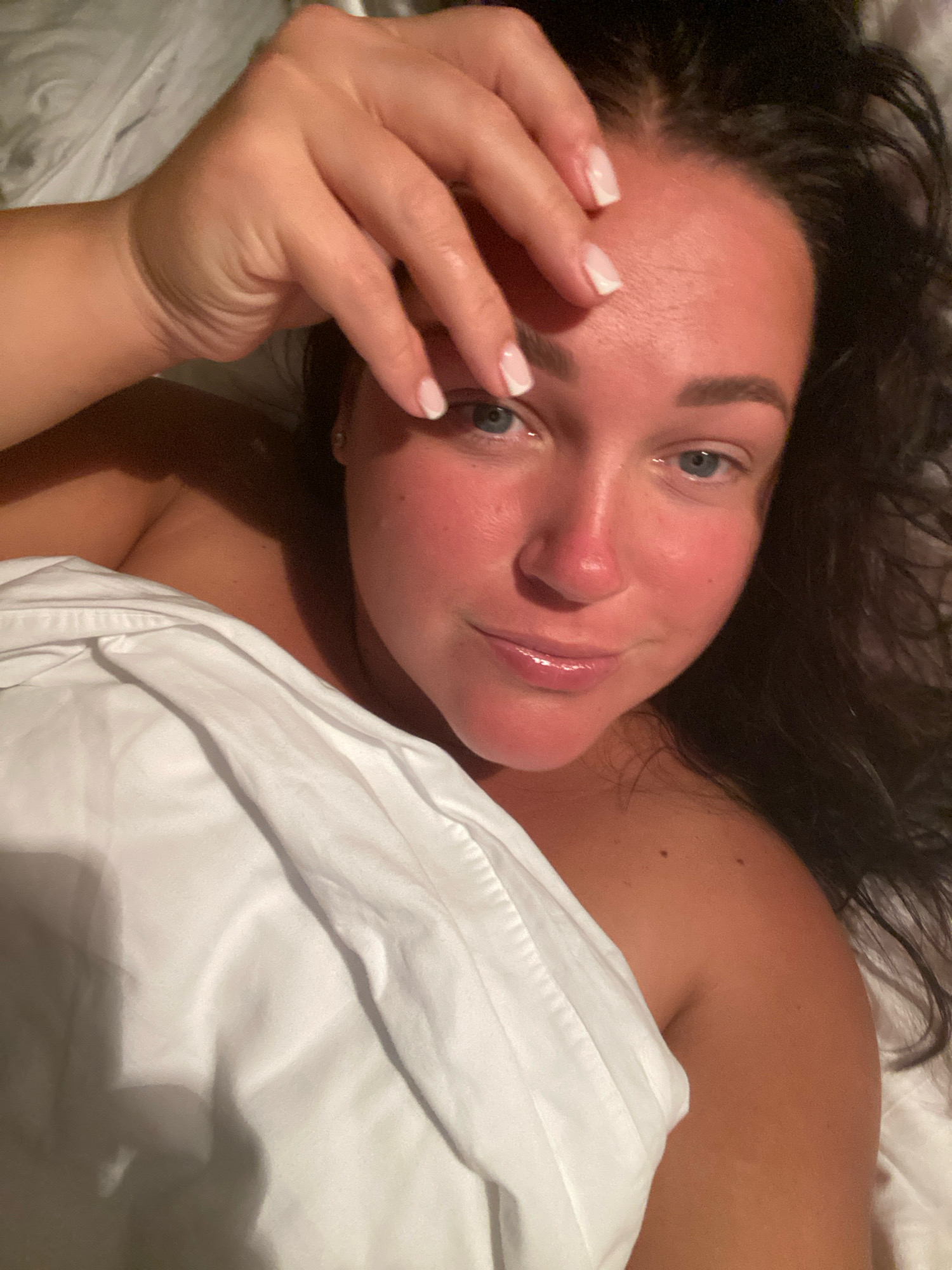 A close-up, low-angle shot features a woman lying down, covered by white bedding. Her right hand rests on her forehead, with manicured fingernails visible. Her dark hair frames her face, and her eyes are open and looking towards the camera. She has red skin on her face and shoulders, likely from sun exposure. The setting appears to be indoors, possibly a bedroom, and the image does not provide any specific details about the city of Nebug, Russia.