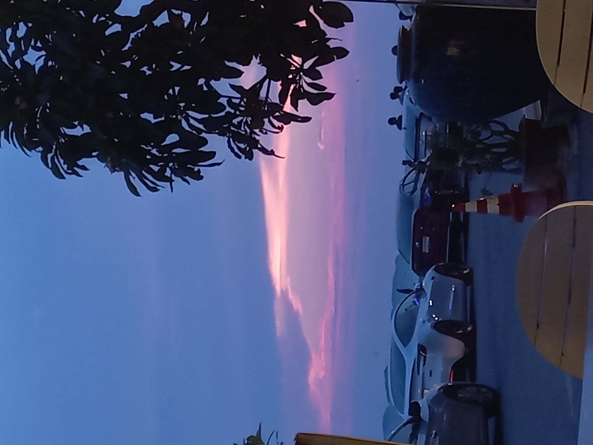 The image depicts a street scene in Pattaya City, Thailand, at dusk. The sky is a gradient of blue and pink, with a vertical band of pink light illuminating the clouds. The foreground is dominated by the silhouette of a tree with dense foliage. Several cars are parked along the right side of the frame, including a white sports car, a dark red car, and a dark sedan. A red and white traffic cone is visible near the parked cars. A large dark-colored jar or container is placed on the sidewalk behind the cars. A portion of a wooden structure, possibly a table or fence, is seen on the extreme right. The overall scene suggests a typical evening in an urban environment in Thailand.
