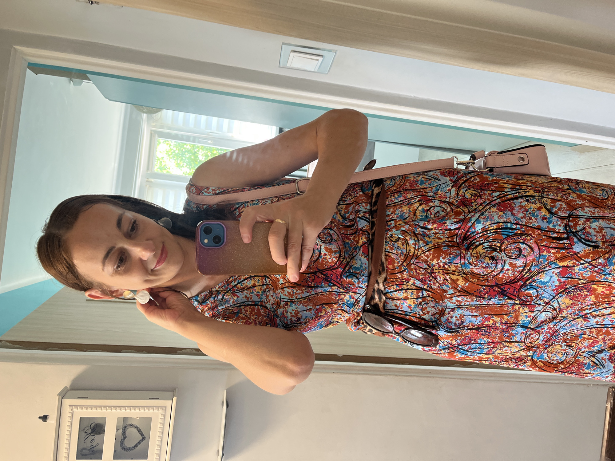 A woman is taking a selfie in a mirror. She has long brown hair and is wearing a colorful, patterned dress. She is also wearing earrings and a ring. A pink purse is slung over her shoulder. A pair of sunglasses with a leopard print frame is tucked into her dress. The background appears to be a hallway or dressing area, with a doorway and a mirror visible. The lighting suggests it is daytime. The woman has a slight smile on her face. There is a decorative framed object with two panes on a shelf below, one pane containing what looks like calligraphy and the other a heart shape.