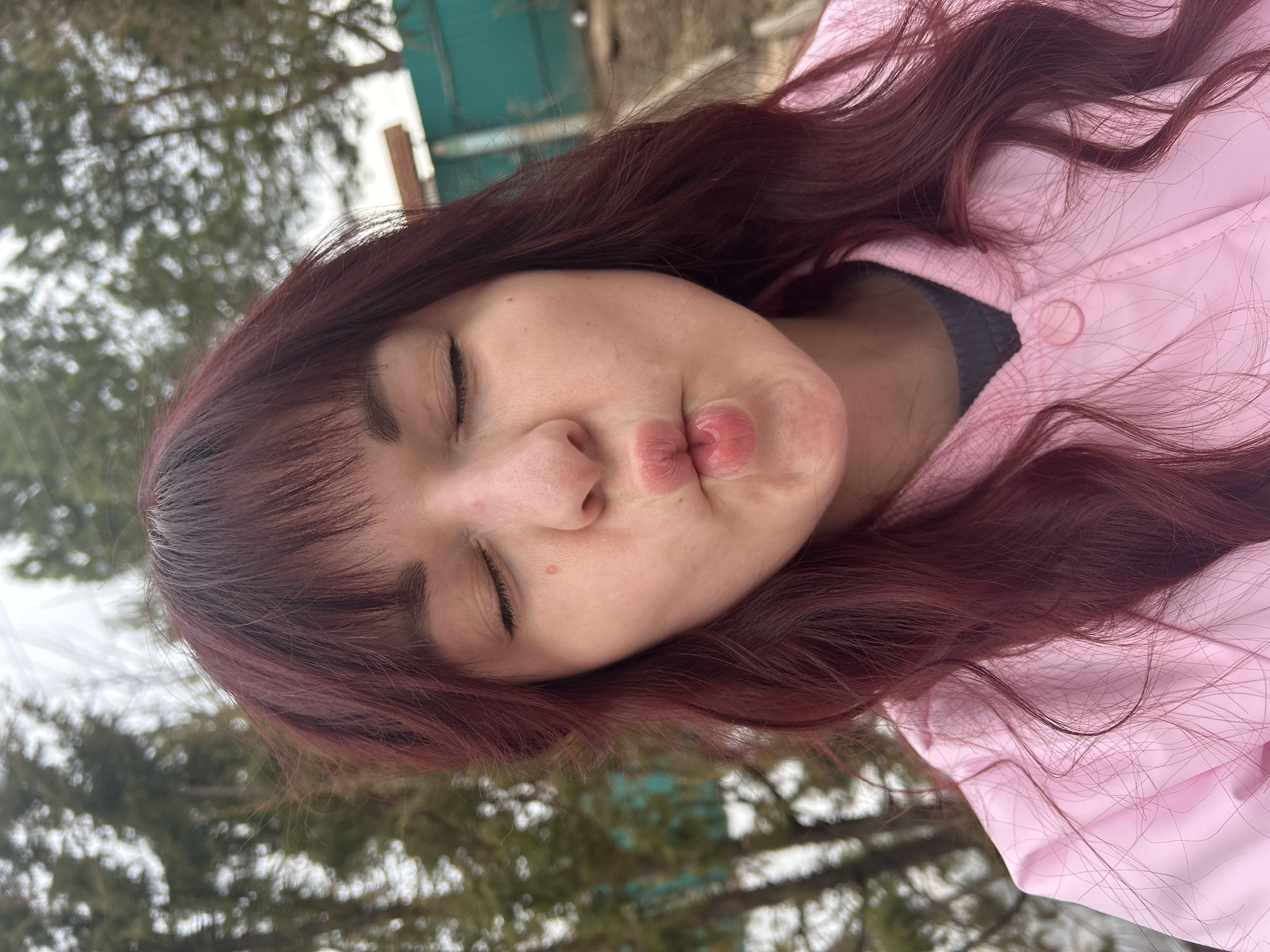 A young person with long, dark red hair is making a kissing face toward the camera. The person has their eyes closed and is wearing a pink jacket. The background appears to be outdoors, with trees and a building partially visible. The location is given as Фоки, Russia.