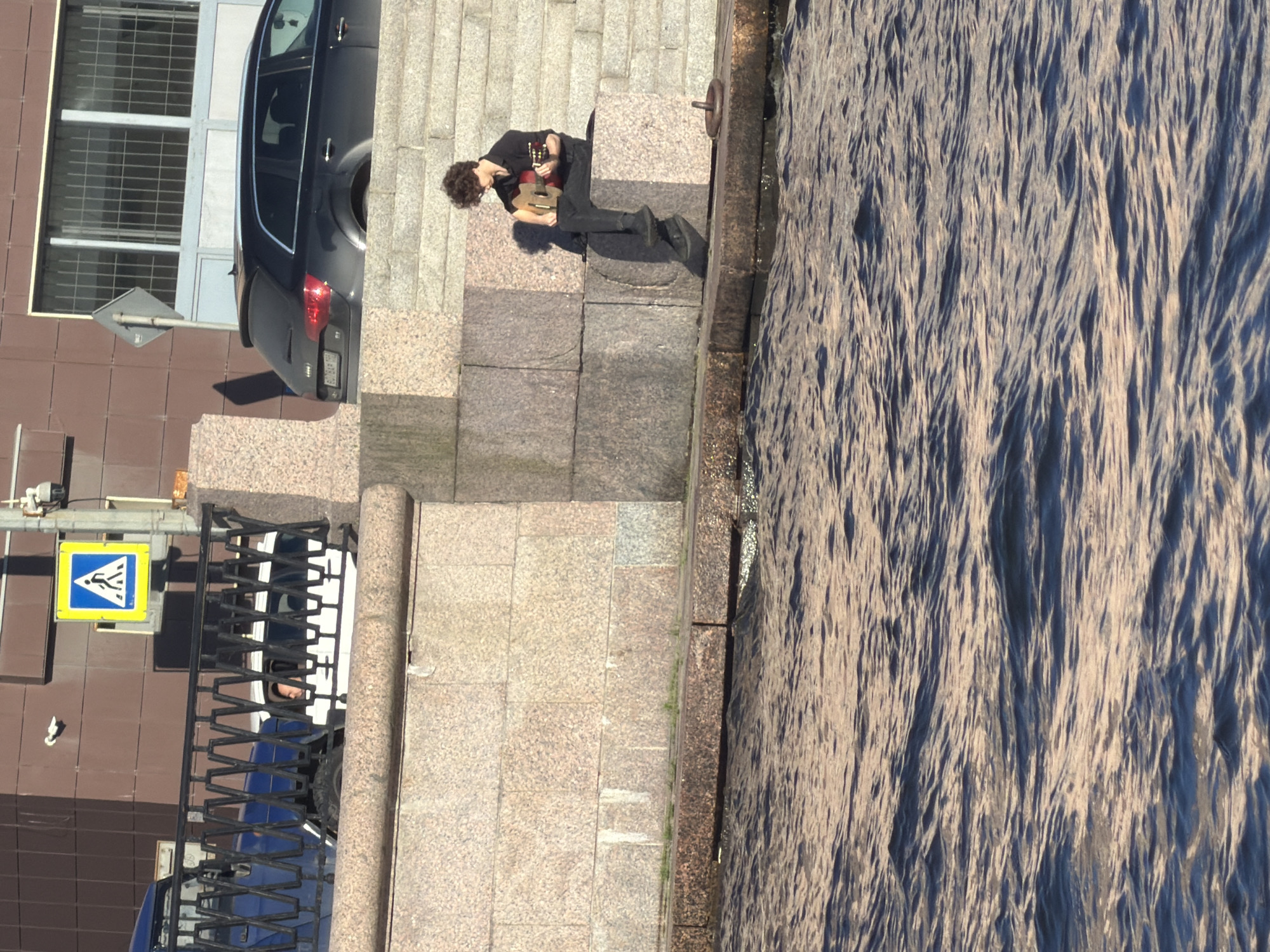 The image depicts a young man with curly hair sitting on the stone steps of a waterfront embankment in Saint Petersburg, Russia, playing a guitar. He is wearing a black shirt and black pants. The embankment is constructed of large, rough-hewn granite blocks. The water in the foreground appears choppy, reflecting the sunlight. In the background, a black Audi station wagon is parked near a building with barred windows. A pedestrian crossing sign is visible on a pole further up the embankment, and a blue and white vehicle is partially obscured by a railing. The scene suggests an outdoor public space, likely along one of the city's many canals or rivers.