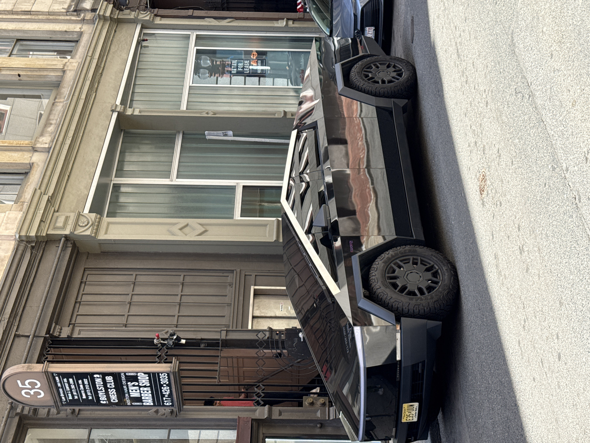 The image shows a black Tesla Cybertruck parked on a city street, close to a building in Boston, Massachusetts. The street is paved with light-colored concrete.
The building is a multi-story structure with architectural details such as cornices and framed windows. A sign in front of the building indicates the address as "35". The sign lists various tenants including law offices, a chess club, and a barber shop.
The Cybertruck has a New Jersey license plate with the alphanumeric "E35 UUN". The vehicle is partially blocking the sidewalk, extending onto the street. The sign “RETAIL FOR LEASE” is visible on a window on the second floor.