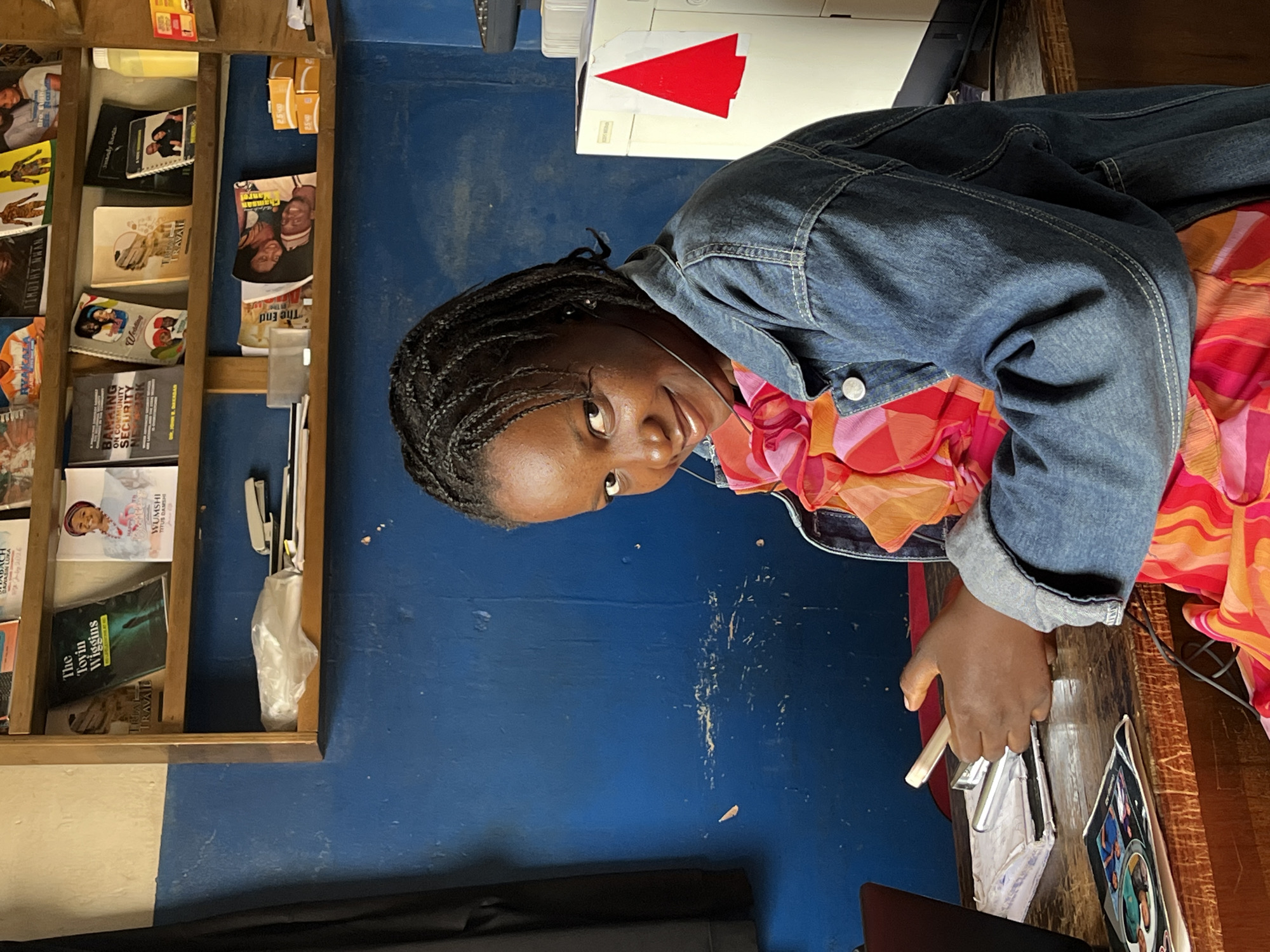 In Jos, Nigeria, a young girl with braided hair is captured in a medium shot, seated at a wooden desk in front of a blue wall. She is looking directly at the camera with a gentle smile, wearing a denim jacket over a vibrant pink and orange patterned dress. Her right hand holds a white mobile phone, and her left hand is resting on the desk, which is cluttered with various items.
On the desk, a laptop is partially visible on the left. In front of the girl, there's a stack of papers or a book, and to the left of it, a stapler. Further to the left, a small, open magazine or booklet displays images of people. A black wire is also visible on the desk.
Behind the girl, a shelf is mounted on the blue wall, filled with books and magazines. Notable titles include "The Toyin Wiggins" and "WUMSHI TITUS DAMSHI." Other items on the shelf include a plastic-wrapped object and what appears to be an office supply.
To the right of the girl, a white office machine, possibly a printer or copier, stands on the desk. A red, triangular sticker is affixed to its front. The overall environment suggests a small office or a study area. The lighting is natural, illuminating the scene without harsh shadows.