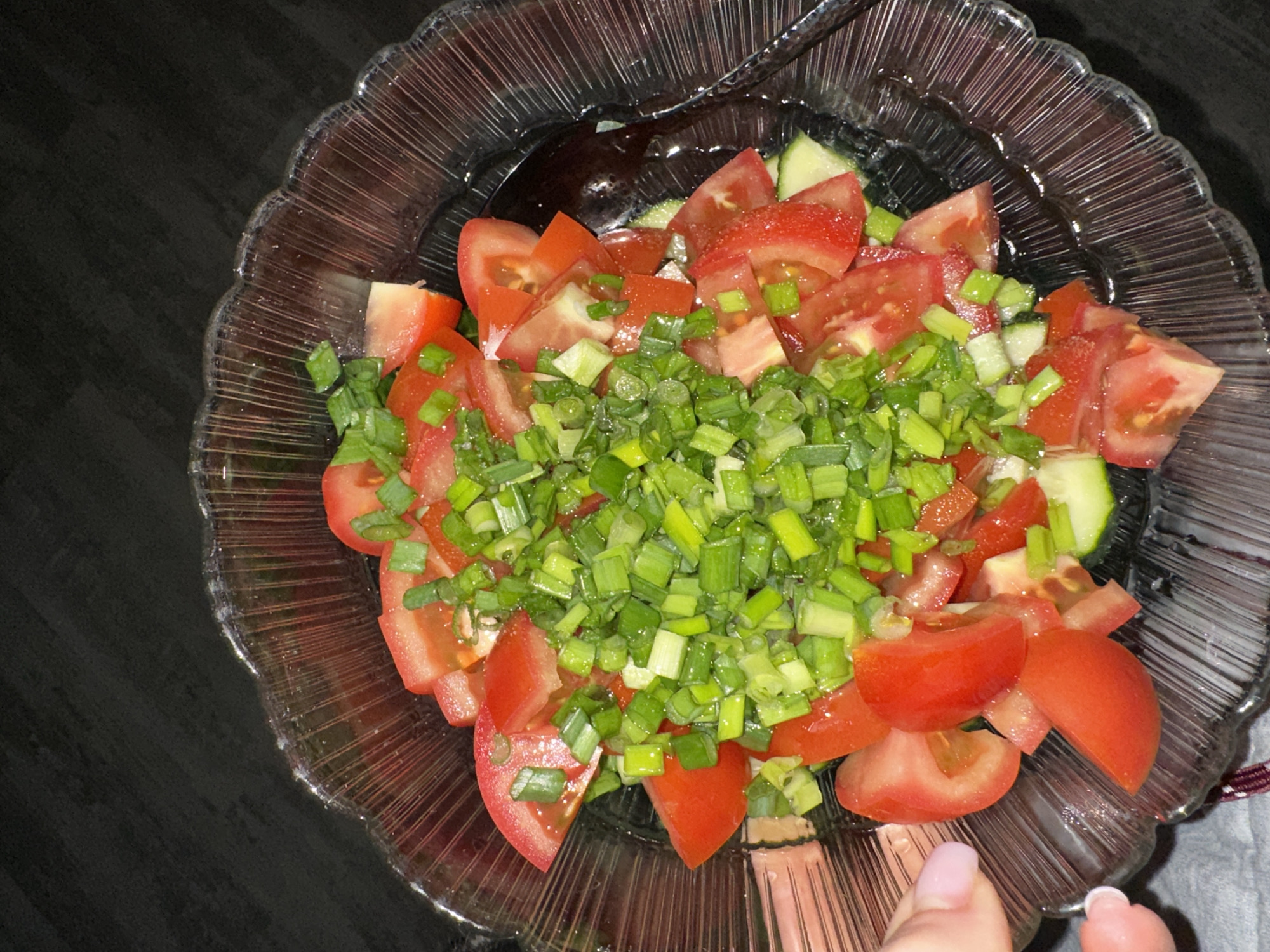 An overhead view captures a clear, fluted glass bowl filled with a fresh salad, resting on a dark, textured surface. The salad primarily consists of irregularly chopped red tomatoes and a generous topping of finely chopped bright green scallions. Diced cucumber pieces are also visible mixed among the tomatoes. A dark serving spoon is partially submerged in the salad on the right side of the bowl. On the lower left, the manicured fingers of a person, with light pink nail polish, gently touch the outer rim of the glass bowl. The scene is well-lit, showing reflections on the glass and the wet ingredients. The image does not depict any identifiable features of the city of Blagoveshchensk, Russia, or any other specific geographical location.