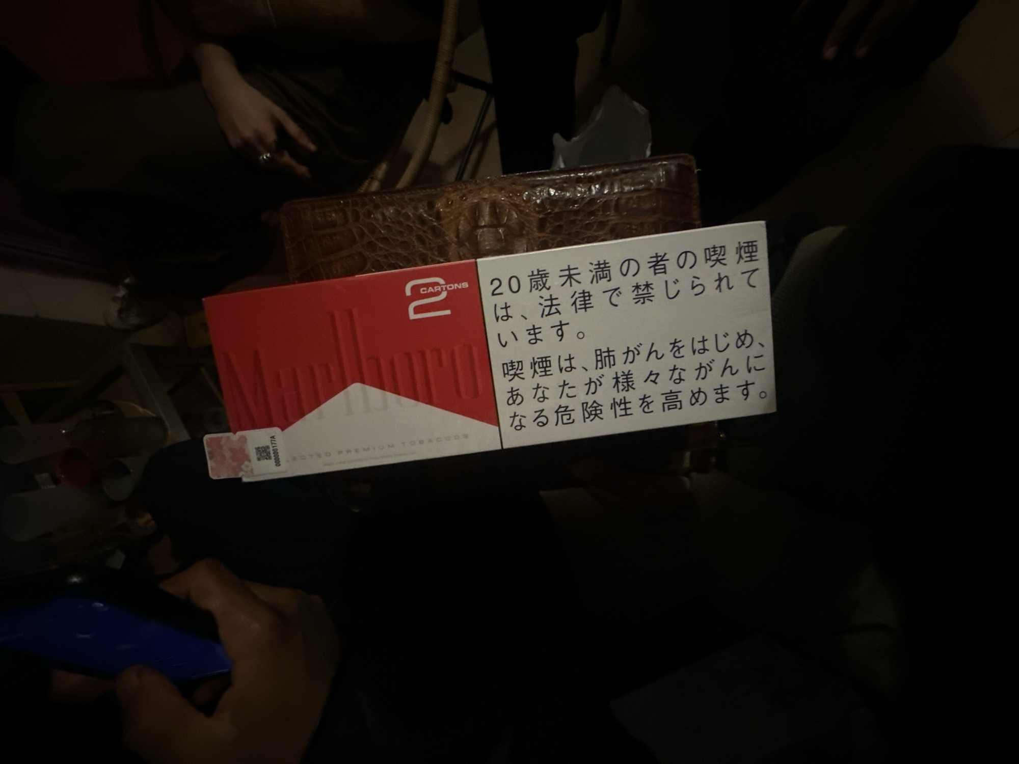 The image shows a pack of Marlboro cigarettes on top of a brown, textured box, possibly a wallet or a tissue box. The cigarette pack has Japanese warning labels printed on its side. A hand holding a smartphone is visible in the lower left corner of the image. The scene appears to be indoors with multiple people present. There is nothing in the image to indicate that it's in Montreuil, France.
