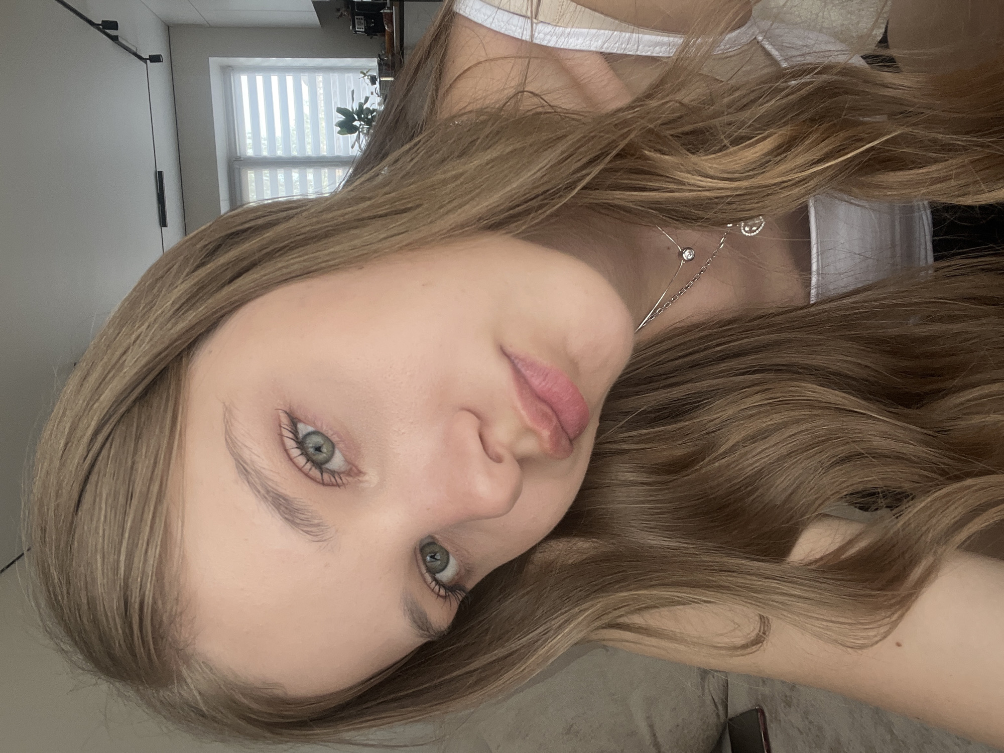 A young woman with light blonde hair and green eyes is shown in a close-up, angled selfie. She is wearing a white tank top and a delicate silver necklace. The background includes a white window with blinds, a dark cabinet, and a glimpse of indoor greenery. There is no definitive evidence to pinpoint the location as Votkinsk, Russia; the setting appears to be indoors, likely a residential setting.