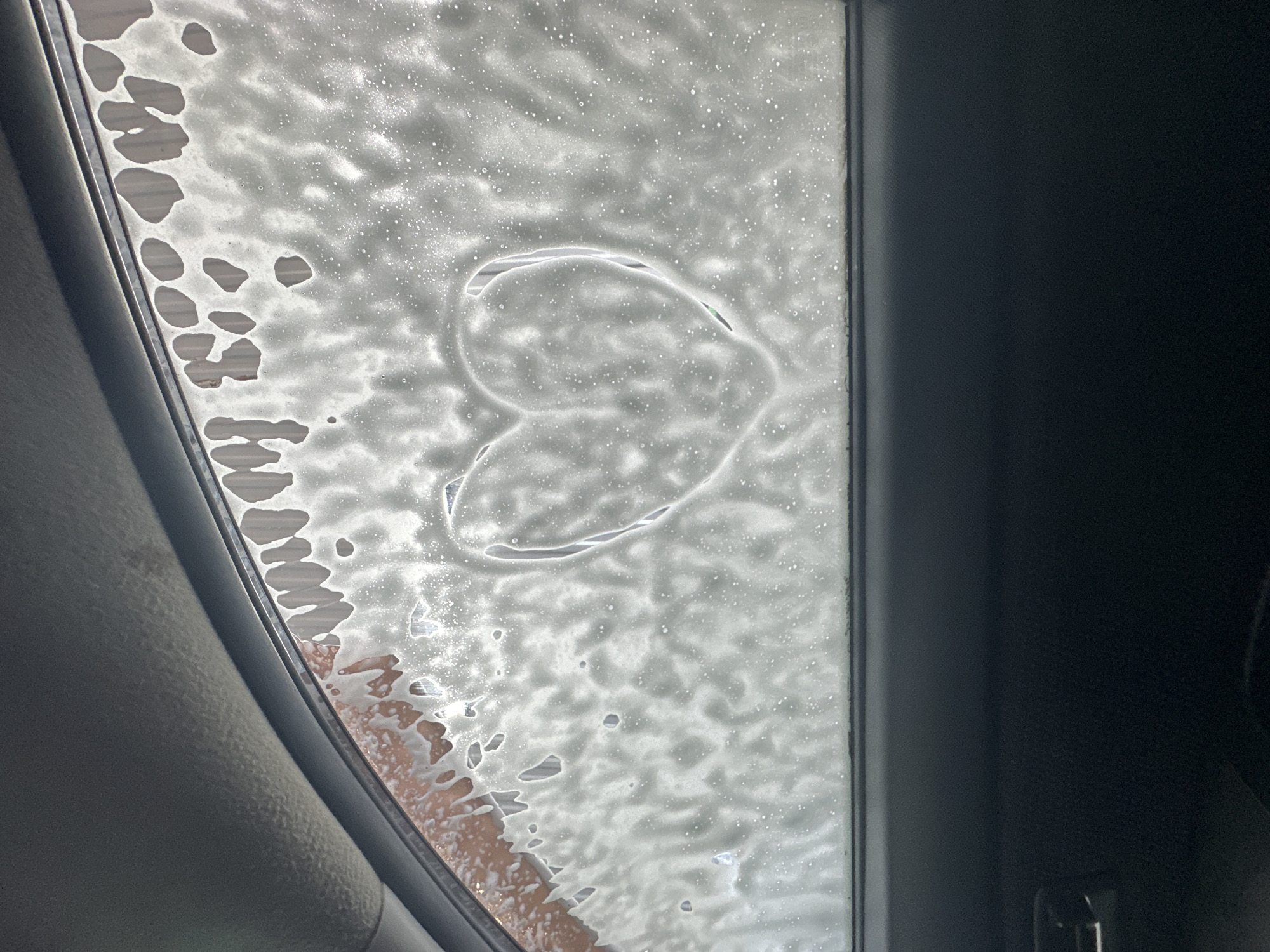 The image shows a heart shape drawn in soap suds on a car window, likely inside a car wash. The view is from inside the vehicle, looking out through the soapy window. The left side of the frame shows the car's interior trim, while the right side shows the door frame. The location (Chaikovsky, Russia) is not evident from the image itself.