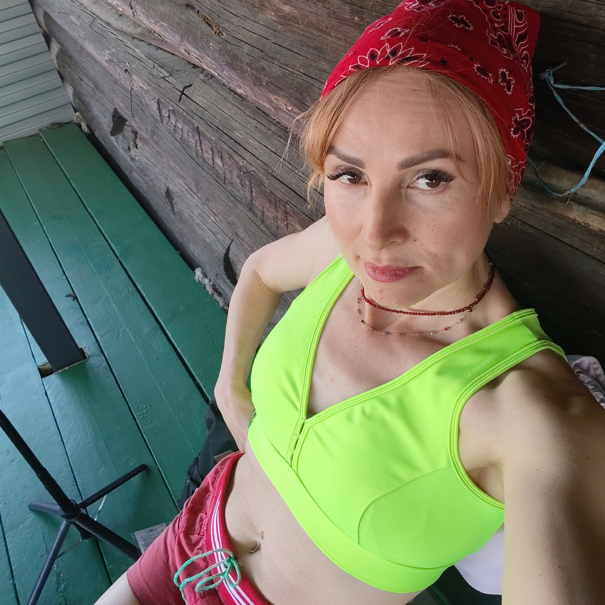 A woman is posing for a selfie. She is wearing a red bandana, a neon green sports bra, and red shorts with white stripes. She has fair skin, light brown hair, and is wearing a red beaded necklace. Her navel is pierced.
The background consists of a wooden structure and a green-painted floor. A black tripod is partially visible in the lower left corner.
There is no evidence to suggest this image was taken in Chaikovsky, Russia.