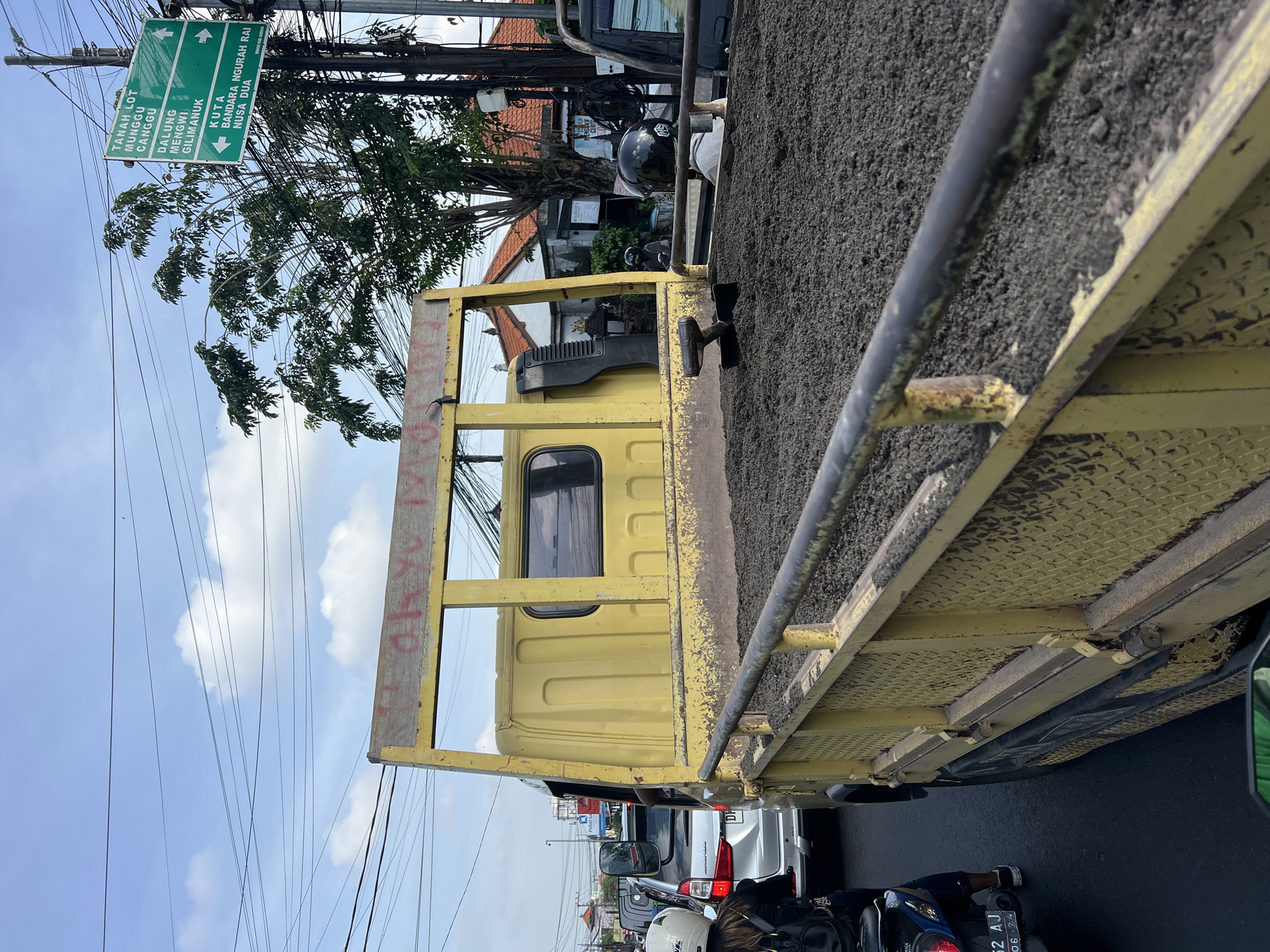 The image shows a yellow truck filled with gravel on a busy street in Denpasar Barat, Indonesia. The truck is likely stopped in traffic, as other cars and motorcycles are visible behind it. A person on a motorcycle is stopped directly behind the truck. Overhead, there are numerous power lines. A green street sign points toward Tanah Lot, Munggu, Canggu, Dalung, Mengwi, Gilimanuk, Kuta, Nusa Dua, and Bandara Ngurah Rai. The sky is partially cloudy.