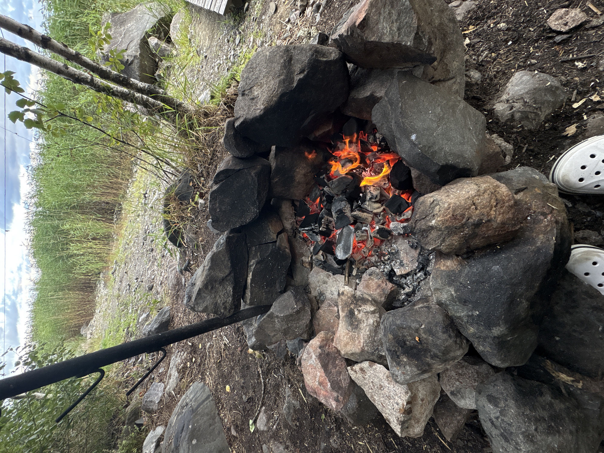 The image depicts a stone-lined fire pit with glowing embers in the center, situated outdoors. Surrounding the fire pit are large, dark rocks and reddish-brown stones, with dry leaves and dirt on the ground. In the background, tall, green reeds or grasses grow abundantly, and a dirt path leads away from the fire pit. Two young birch trees with striped bark are visible to the right of the fire pit, with some branches extending over the scene. The sky above is cloudy. In the foreground, two white Croc-style shoes are visible, indicating human presence. A black metal pole with two curved hooks stands to the left of the fire pit, possibly for cooking or hanging items. To the far right, a wooden structure, possibly part of a bench or boardwalk, is partially visible. The overall scene suggests a natural, perhaps slightly wild, outdoor setting, consistent with a location like Rautalakhti, Russia, where such rustic arrangements for campfires might be found.