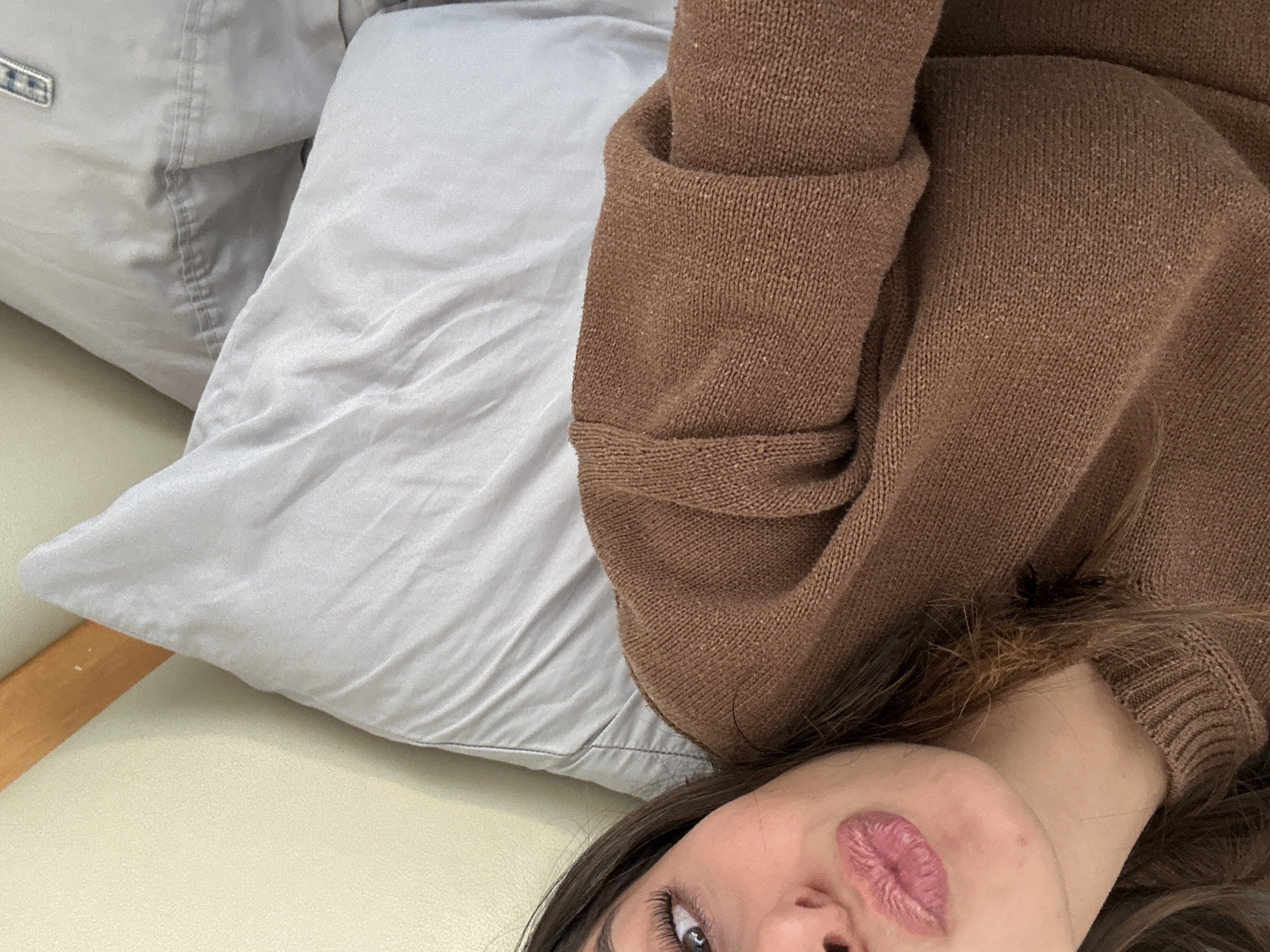 A close-up, eye-level shot depicts a person lying down with their face partially visible in the bottom right corner of the frame. The person has brown hair and pink lipstick. They are wearing a brown sweater. The person is resting on a light grey pillow, which is on top of a beige surface. A corner of another grey pillow is seen in the top left. The location and city are unknown.