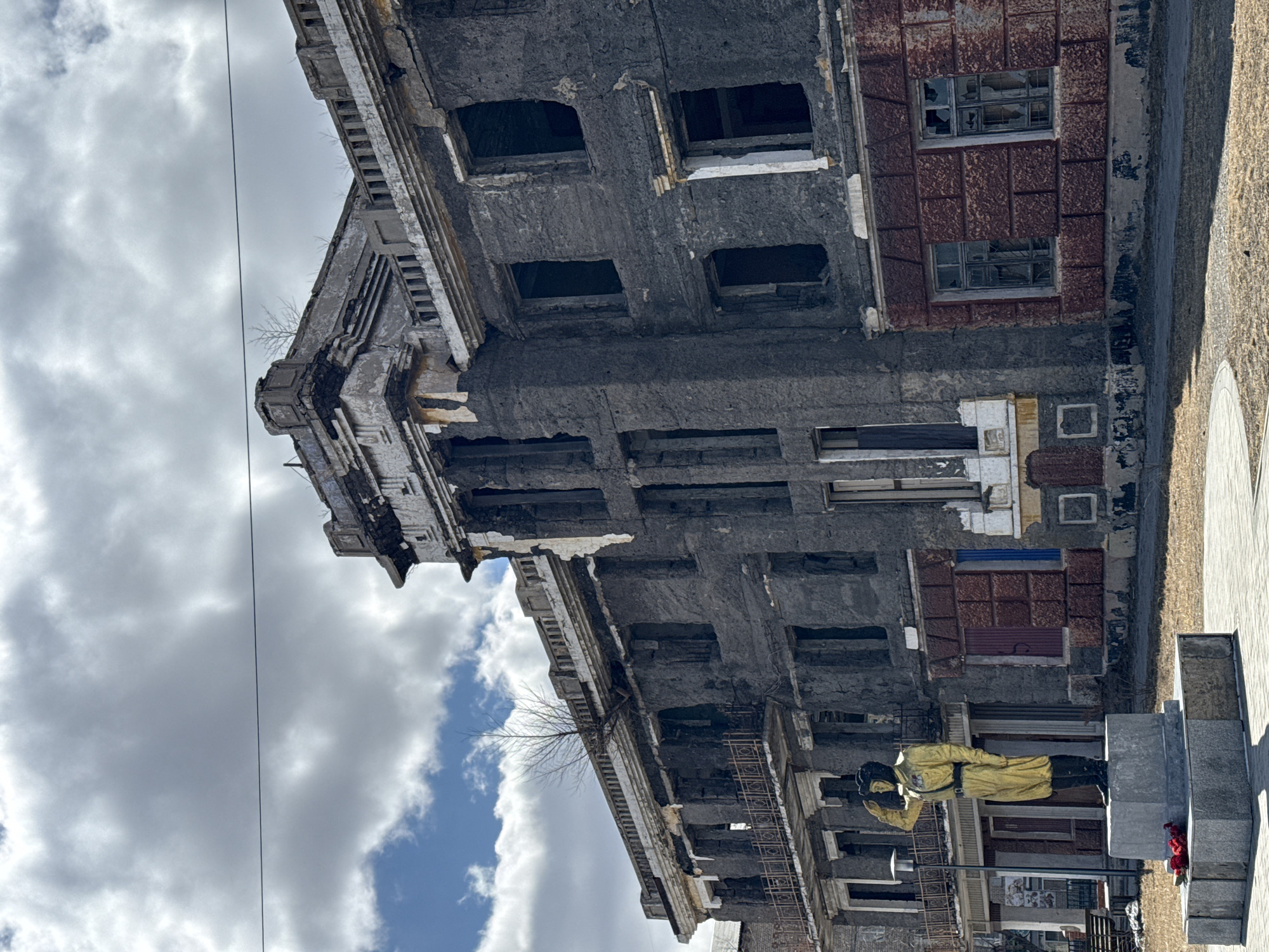 The image shows a dilapidated three-story building in Borzya, Russia. The building's facade is heavily weathered and crumbling, with missing window panes and exposed structural elements. Some sections have a reddish-brown brick-like texture. A statue of a person in a yellow outfit stands in front of the building on a stone pedestal, with red flowers nearby. Overhead, there are clouds and a utility line.