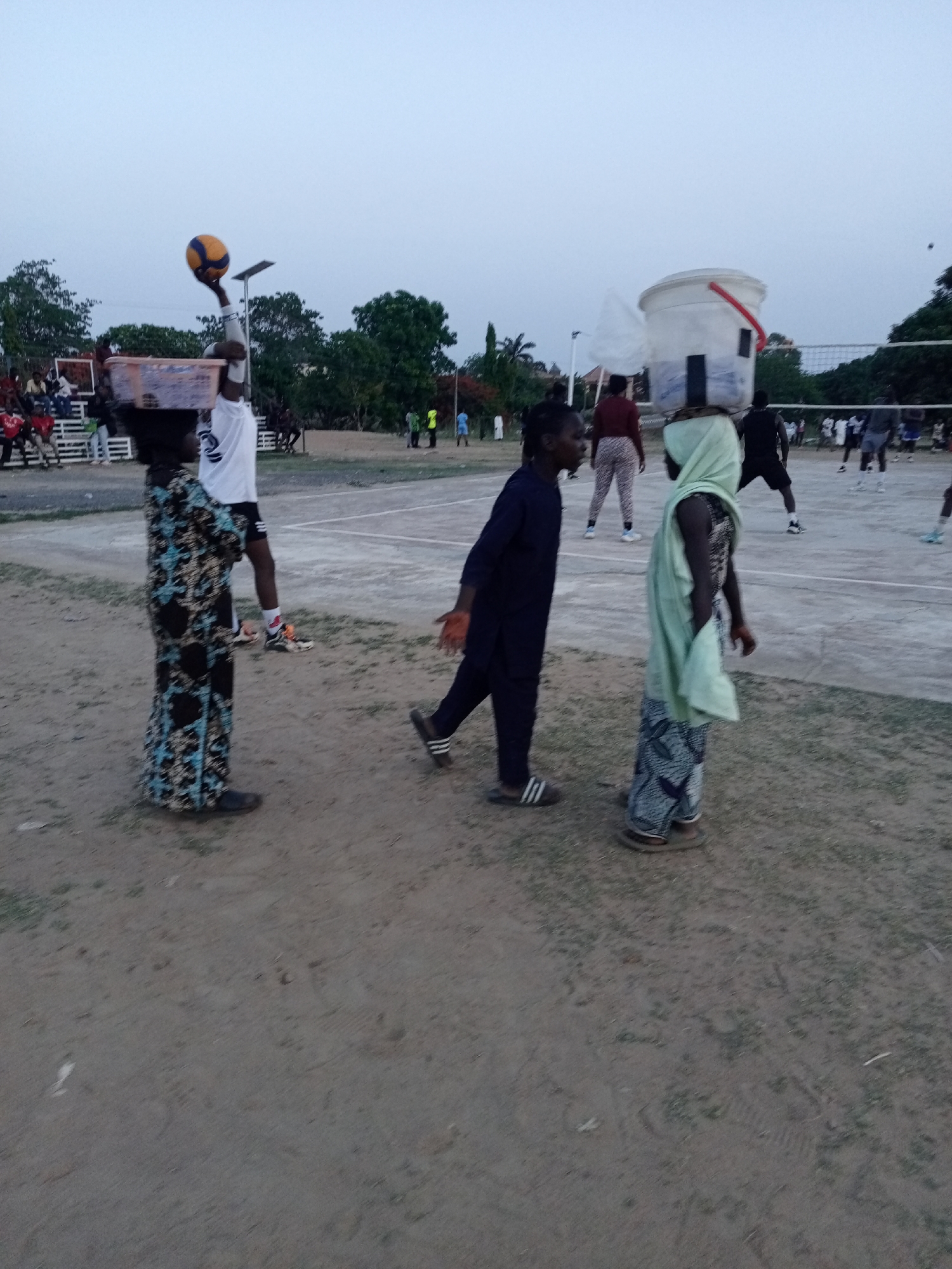 This image captures an outdoor scene at dusk in Keffi, Nigeria, depicting a blend of sports and daily activities. In the foreground, three individuals are prominent. On the left, a woman wearing a long, patterned dress balances a rectangular basket on her head. Next to her, a man in a dark blue tunic and trousers with striped sandals walks towards the right. To his right, another woman in a light green headscarf and a patterned skirt carries a large white bucket on her head.
In the mid-ground, a vibrant volleyball game is in progress on a paved court. Several players are visible, with one player in a white shirt and shorts jumping to hit a yellow and blue volleyball. A white volleyball net stretches across the court, dividing the players.
The background shows spectators seated on bleachers to the left, and other people gathered around the court. Lush green trees border the playing area, and solar-powered streetlights are visible, indicating the time of day is early evening or twilight. The ground in the foreground is a mix of dirt and sparse grass. The sky above is a pale, clear blue.
Visible details include the distinct yellow and blue pattern of the volleyball, the striped design on the sandals worn by the man in dark blue, and a number "2" on the white shirt of the volleyball player. The scene portrays a communal atmosphere with both athletic engagement and people going about their routines.