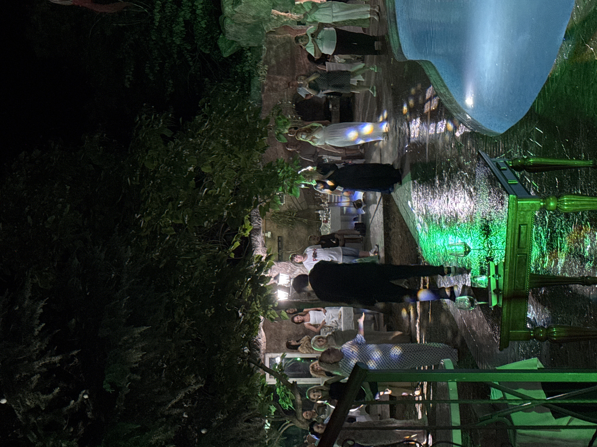 The image depicts an outdoor evening gathering at a venue with a swimming pool. A group of approximately twenty individuals, both male and female, are present. They are dressed in casual attire. Some are standing and conversing, while others appear to be observing an event. The scene is illuminated by artificial lighting, casting reflections on the wet ground, possibly from recent rain or a sprinkler system. The background features trees, a building with windows, and decorative rock formations. A table with wine glasses and a small plate is in the foreground. The overall impression is that of a social event or party taking place in a garden or courtyard setting. The location is identified as Sredets, Bulgaria.